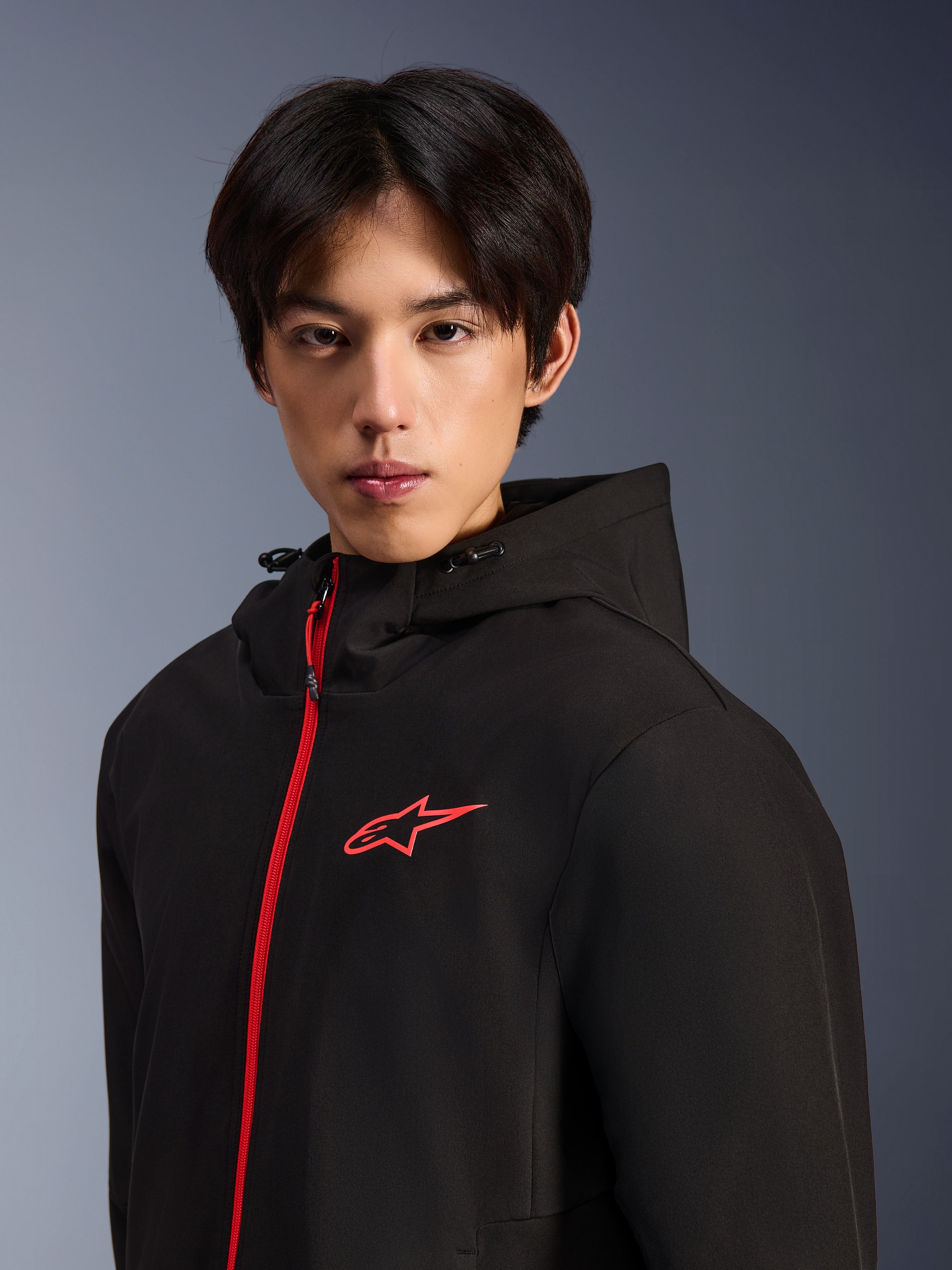 Alpinestars Radium Tech Hoodie, giacca da moto urbana, nero e rosso acceso, modello che indossa una felpa tecnica con cappuccio con cerniera frontale e logo sul petto in rosso acceso, design moderno per la guida urbana