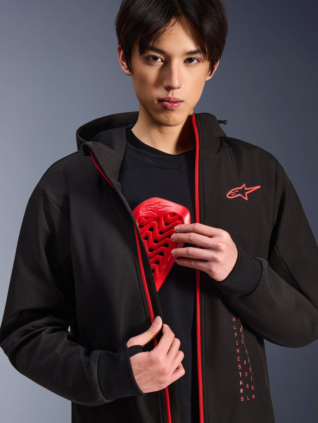 Alpinestars Radium Tech Hoodie, giacca da moto urbana, nero e rosso brillante, indossata da un modello che mostra un inserto protettivo per il torace rosso, con cerniere rosso brillante e branding Alpinestars verticale, guscio tecnico antivento per il commuting urbano