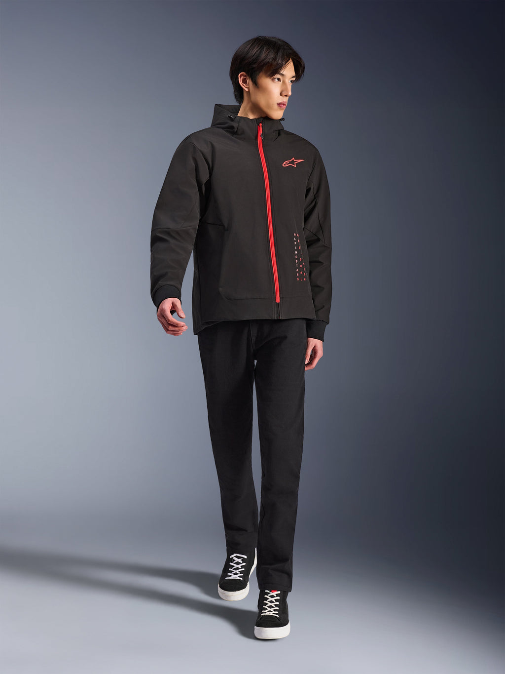 Alpinestars Radium Tech Hoodie in Black Bright Red, giacca da moto urbana indossata da un modello, caratterizzata da una cerniera frontale rossa e logo sul petto, abbinata a pantaloni neri e sneaker per un look da commuting moderno.