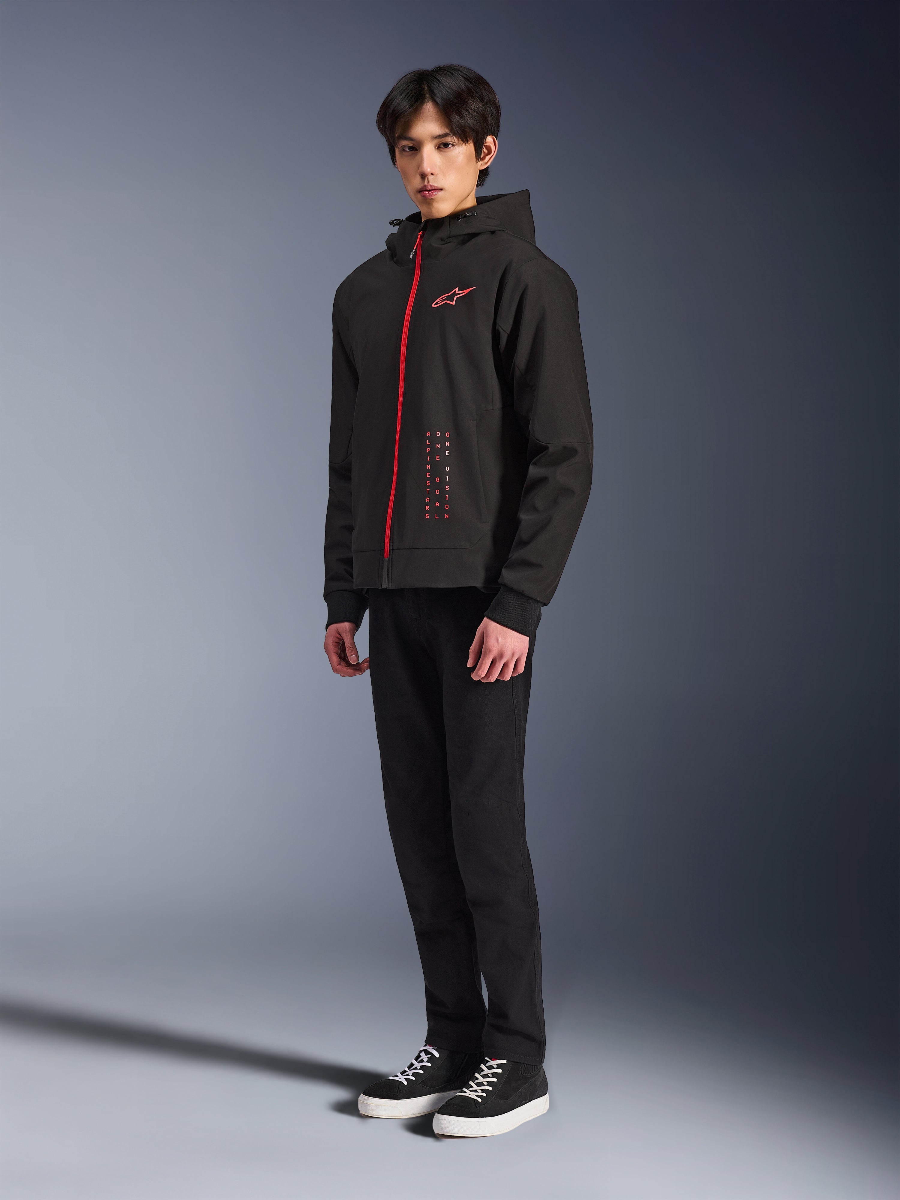Alpinestars Radium Tech Hoodie, Nero Rosso Brillante, felpa tecnica per la guida urbana con cerniera frontale rossa e logo sul petto, indossata da un modello con pantaloni neri e sneakers nere con suola bianca, scatto in studio a figura intera