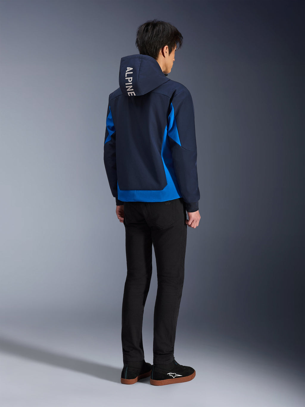 Alpinestars Radium Tech Hoodie, Night Navy e Royal Blue, vista posteriore del modello che indossa la felpa tecnica con scritta 'ALPINE' bianca sul cappuccio, abbinata a pantaloni e scarpe neri, abbigliamento da moto urbano