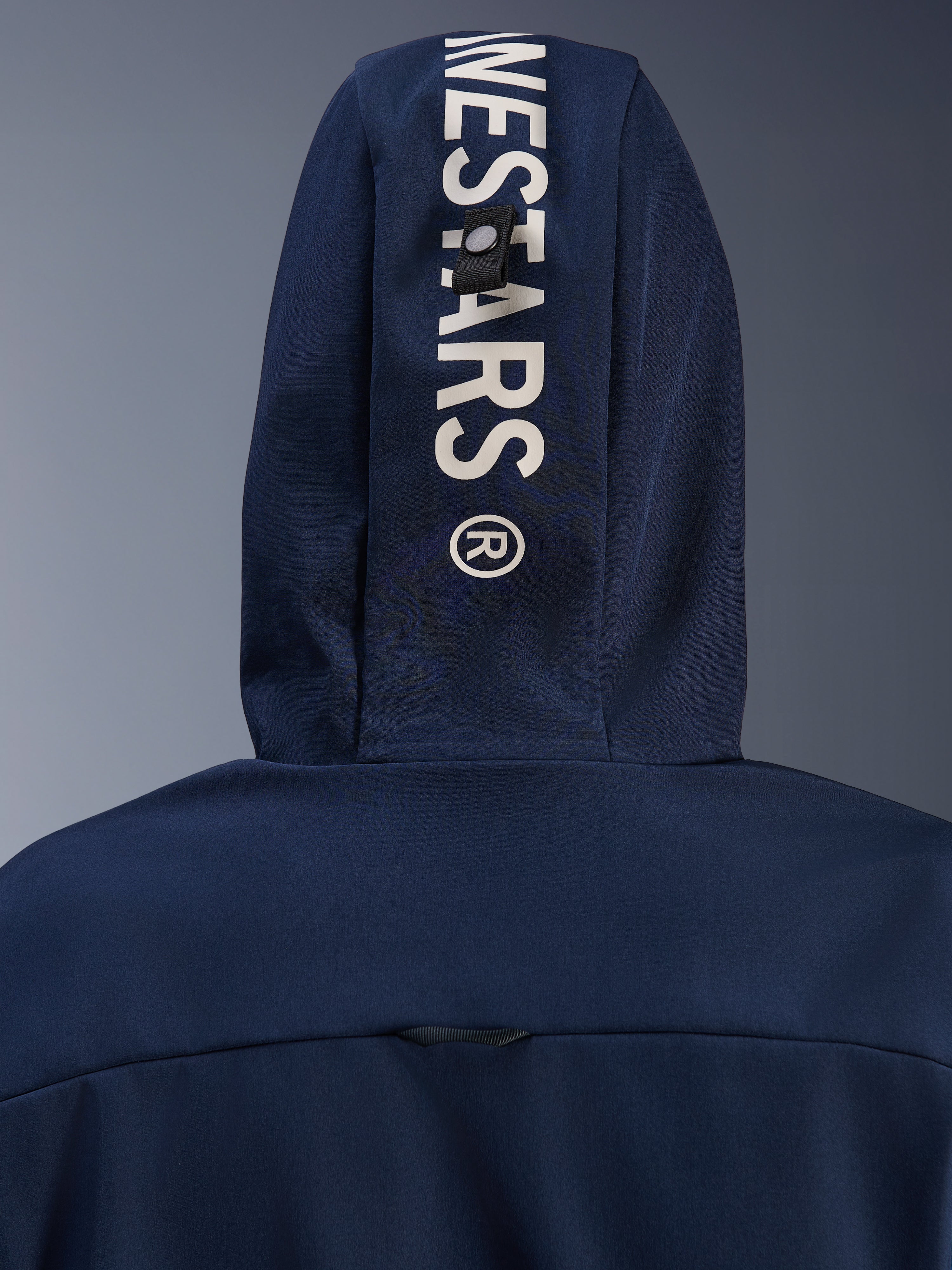 Alpinestars Radium Tech Hoodie in Night Navy Royal Blue, primo piano del cappuccio posteriore con branding Alpinestars verticale bianco e sistema di fissaggio ad asola, abbigliamento tecnico da moto urbano.