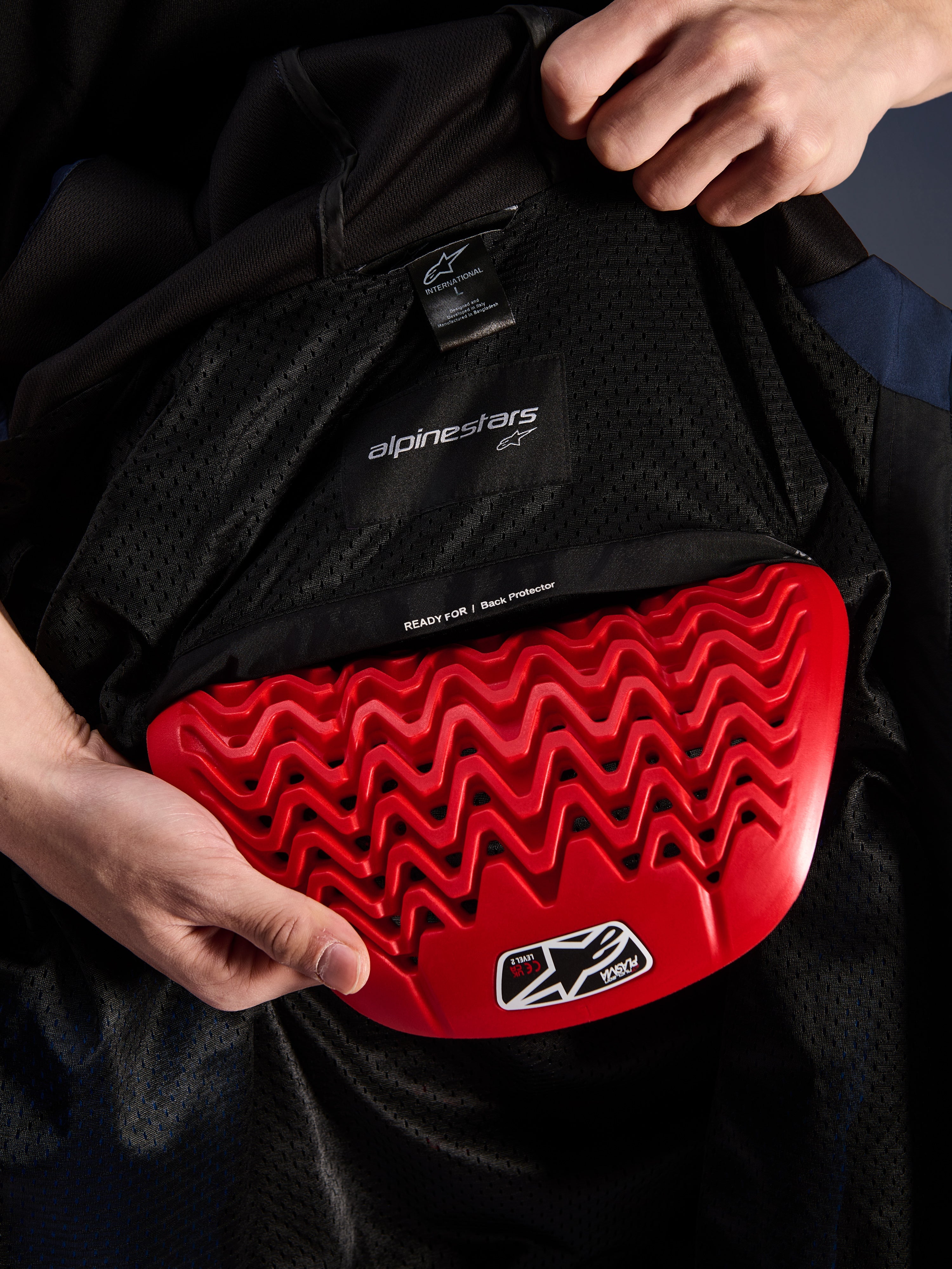 Dettaglio interno della felpa Alpinestars Radium Tech Hoodie, paraschiena rosso Nucleon Plasma inserito nella tasca dedicata, fodera in mesh nera con scritta 'READY FOR | Back Protector', design a nido d'ape ondulato, caratteristiche protettive per giacca da moto urbana tecnica