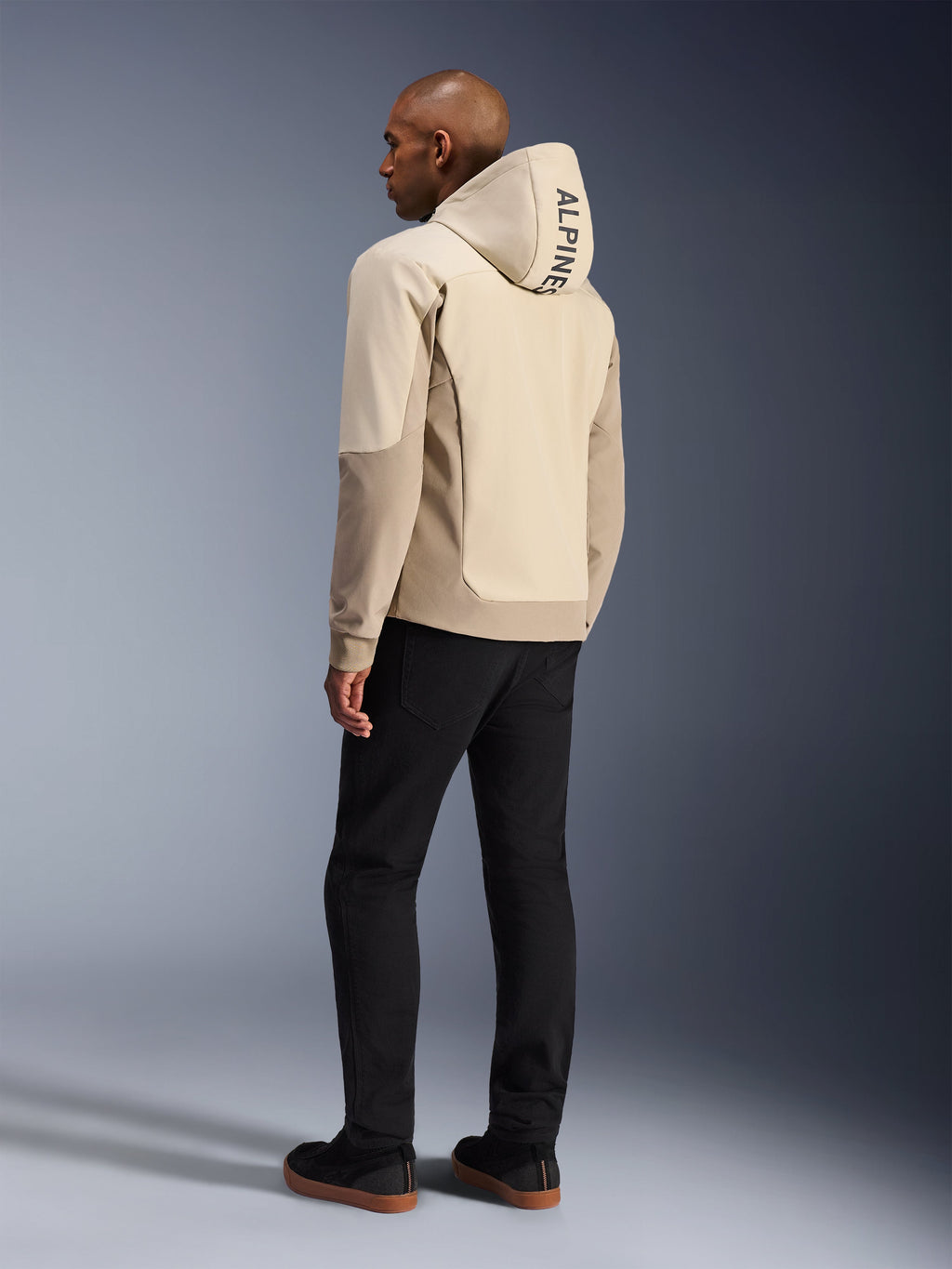 Alpinestars Radium Tech Hoodie in Mastic Aluminum, giacca da moto urbana, vista posteriore su modello con cappuccio alzato, logo ALPINESTARS verticale sul cappuccio, abbinata a pantaloni neri, moderna costruzione in tessuto tecnico per la protezione nella guida urbana.