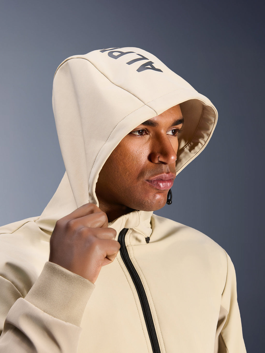 Alpinestars Radium Tech Hoodie in Mastic Aluminum, primo piano di un uomo con il cappuccio sollevato, logo Alpinestars sulla parte superiore, giacca da moto urbana con guscio tecnico antivento, estetica streetwear moderna per la guida in città