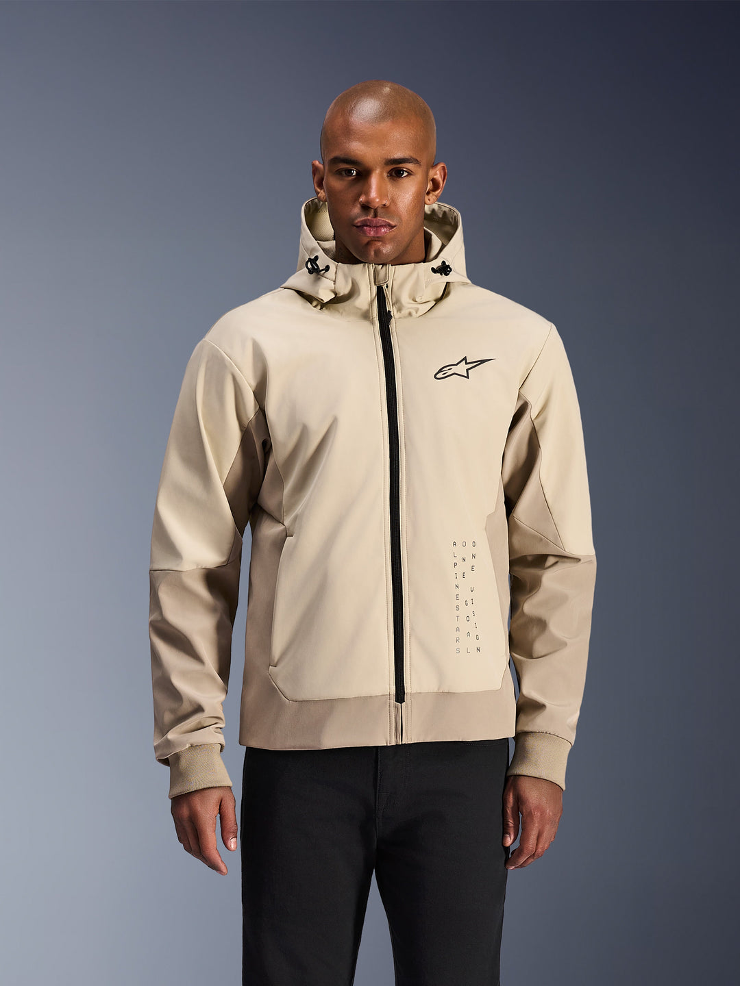 Alpinestars Radium Tech Hoodie in Mastic Aluminum, giacca da moto urbana beige chiaro e tan, cerniera anteriore nera e piccolo logo sul petto, indossata da un uomo con pantaloni neri, guscio tecnico antivento dallo stile streetwear contemporaneo