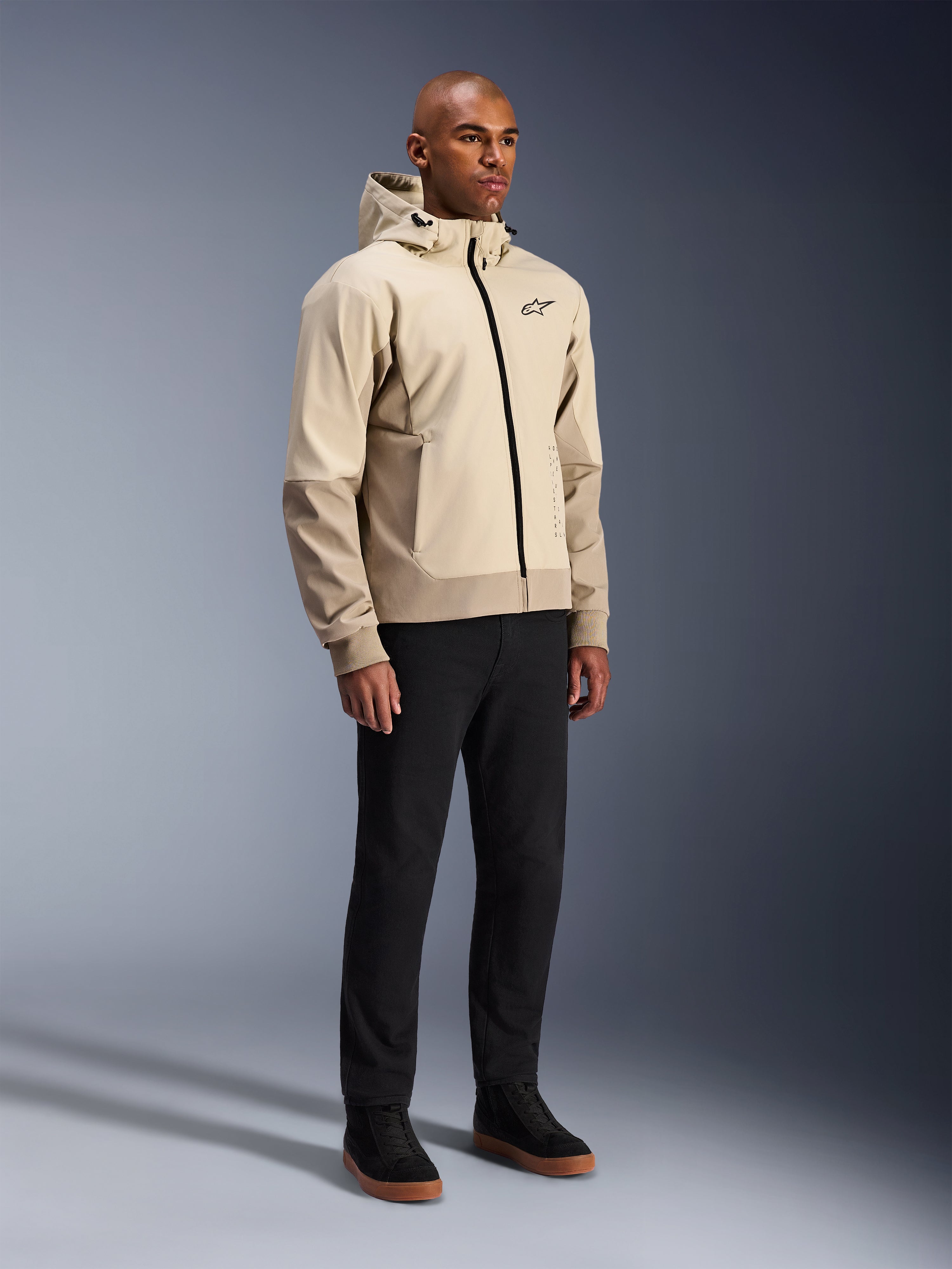 Alpinestars Radium Tech Hoodie, giacca da moto urbana, beige chiaro Mastic Aluminum, indossata con pantaloni neri e sneaker alte nere, dotata di guscio tecnico antivento, cerniera frontale e cappuccio rimovibile, design streetwear moderno per la guida urbana