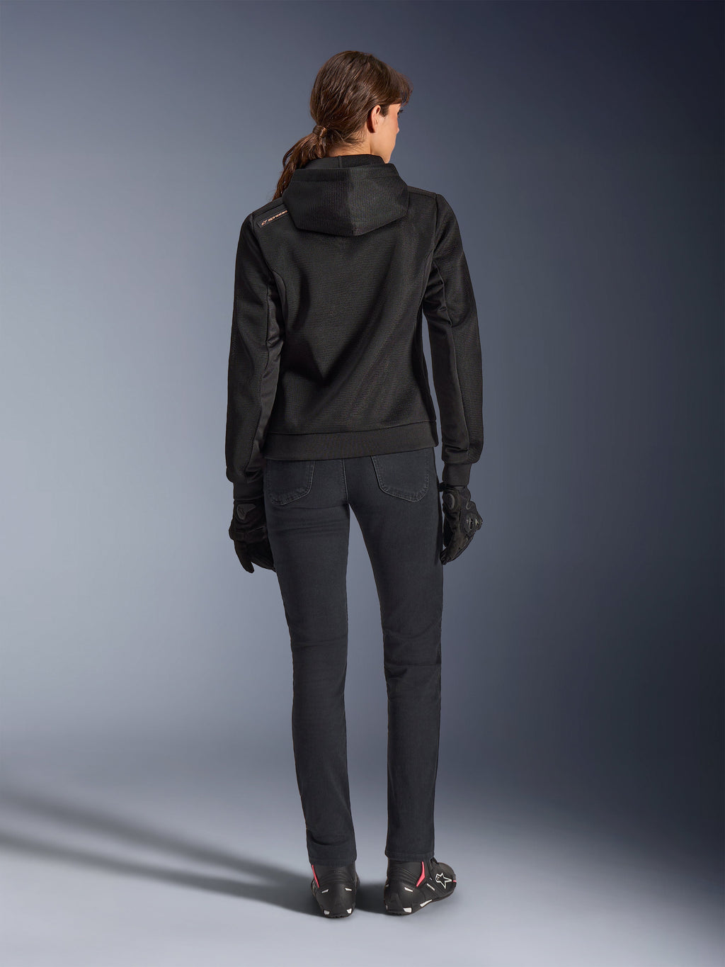 Alpinestars Stella Chrome Superairflow Hoodie, felpa tecnica da moto da donna, mesh 3D nera con piccolo logo arancione sulla spalla, vista posteriore, indossata con pantaloni da moto neri, guanti e stivali neri, progettata per la guida urbana sportiva