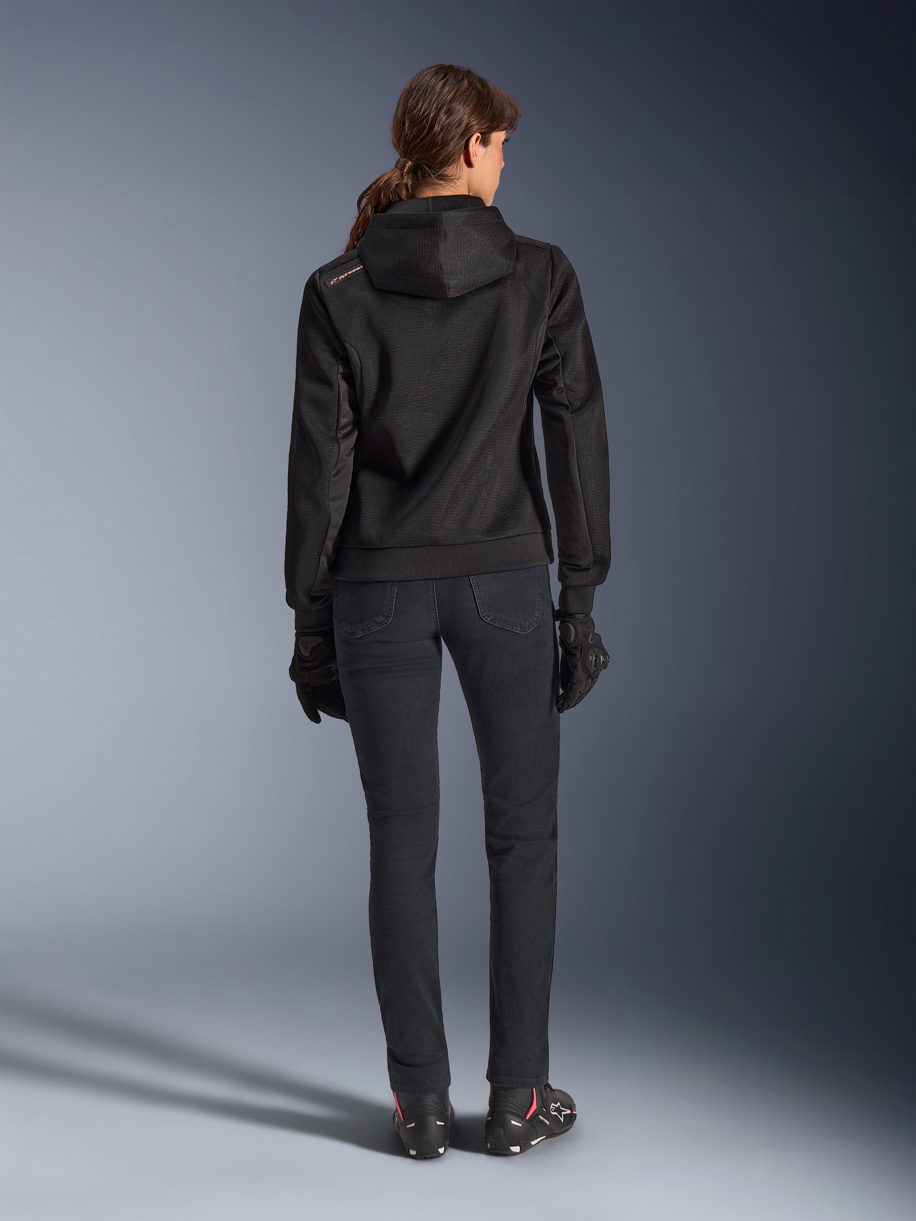 Alpinestars Stella Chrome Superairflow Hoodie, felpa tecnica da moto da donna, mesh 3D nera con piccolo logo arancione sulla spalla, vista posteriore, indossata con pantaloni da moto neri, guanti e stivali neri, progettata per la guida urbana sportiva