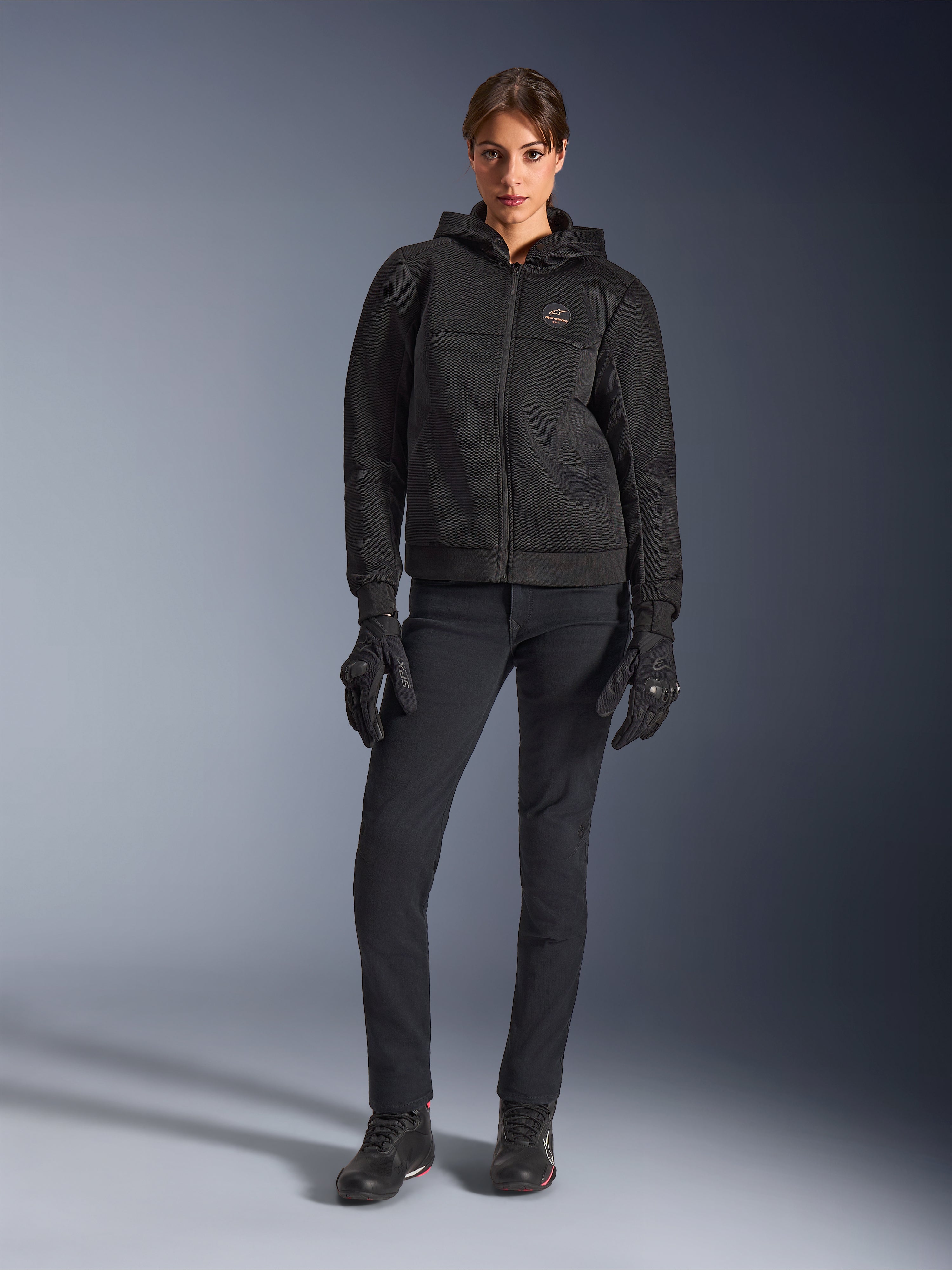 Modella che indossa la Alpinestars Stella Chrome Superairflow Hoodie in nero, abbinata a pantaloni tecnici e guanti da guida neri, caratterizzata da una costruzione in mesh 3D e logo sul petto color rame, abbigliamento da moto urban sport per donna