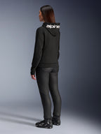Donna che indossa la felpa Alpinestars Stella Chrome V2 Sport Hoodie, vista posteriore, felpa da moto nera con logo Alpinestars bianco sul cappuccio, abbinata a pantaloni tecnici da guida neri e scarpe da moto nere, abbigliamento casual per il pendolarismo urbano