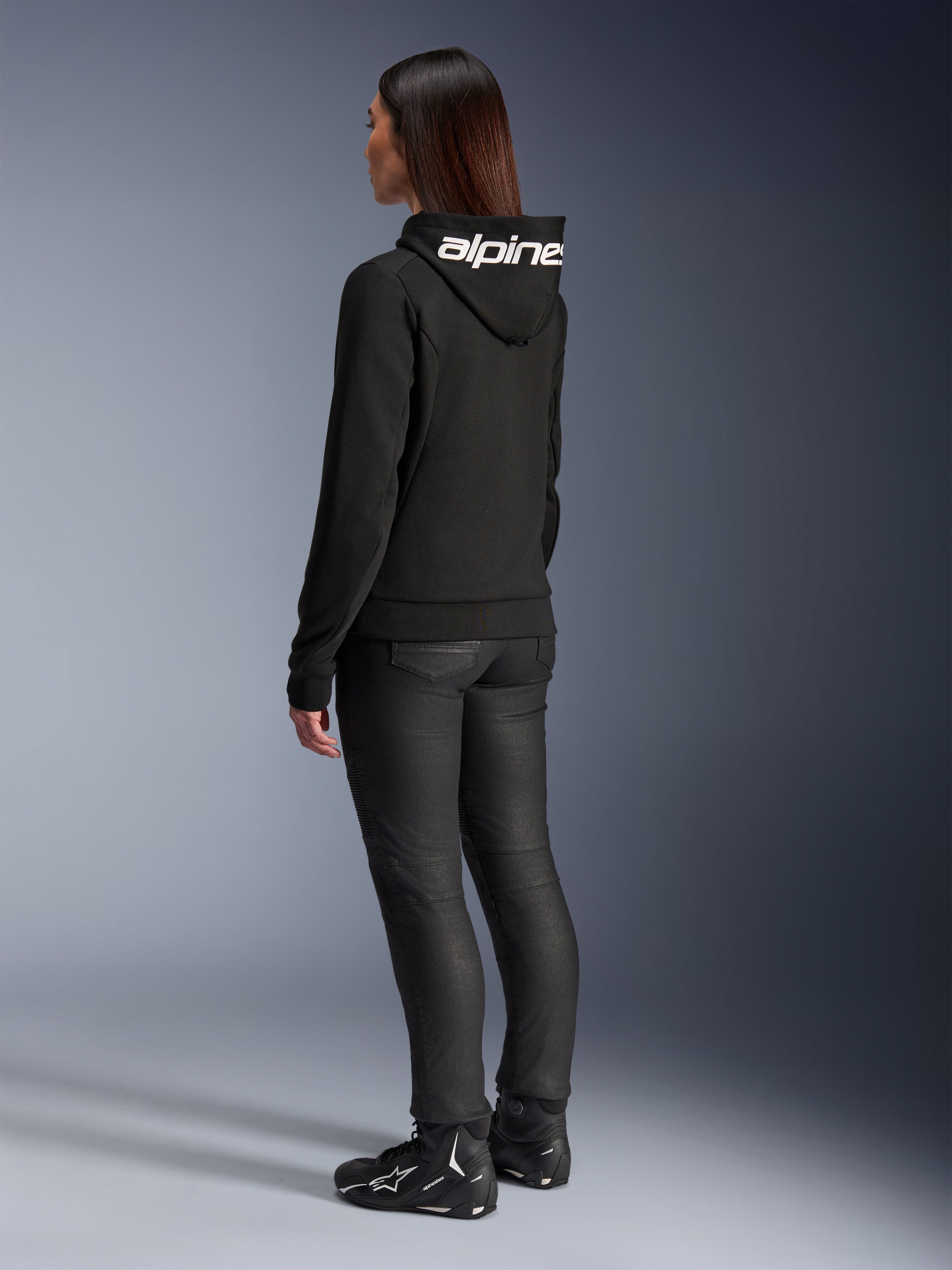 Donna che indossa la felpa Alpinestars Stella Chrome V2 Sport Hoodie, vista posteriore, felpa da moto nera con logo Alpinestars bianco sul cappuccio, abbinata a pantaloni tecnici da guida neri e scarpe da moto nere, abbigliamento casual per il pendolarismo urbano