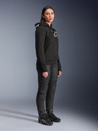 Donna che indossa la felpa Alpinestars Stella Chrome V2 Sport Hoodie in nero, con logo Alpinestars bianco, abbinata a pantaloni e stivali da moto neri, abbigliamento urban da donna con vestibilità atletica casual