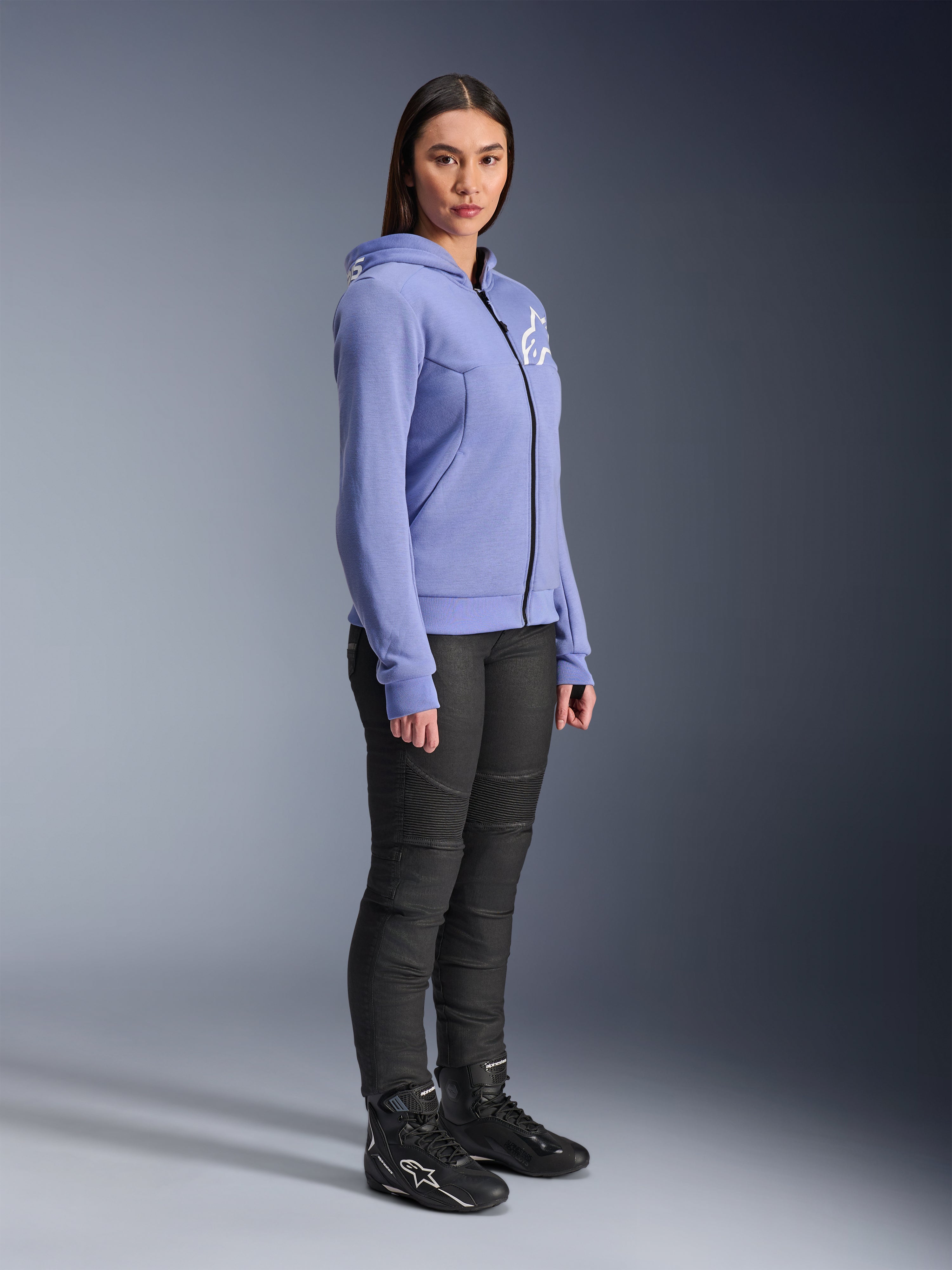 Alpinestars Stella Chrome V2 Sport Hoodie da donna, felpa da moto blu-viola con logo Alpinestars bianco sul petto, indossata da una modella con pantaloni da moto e stivali neri, abbigliamento urban riding per donna
