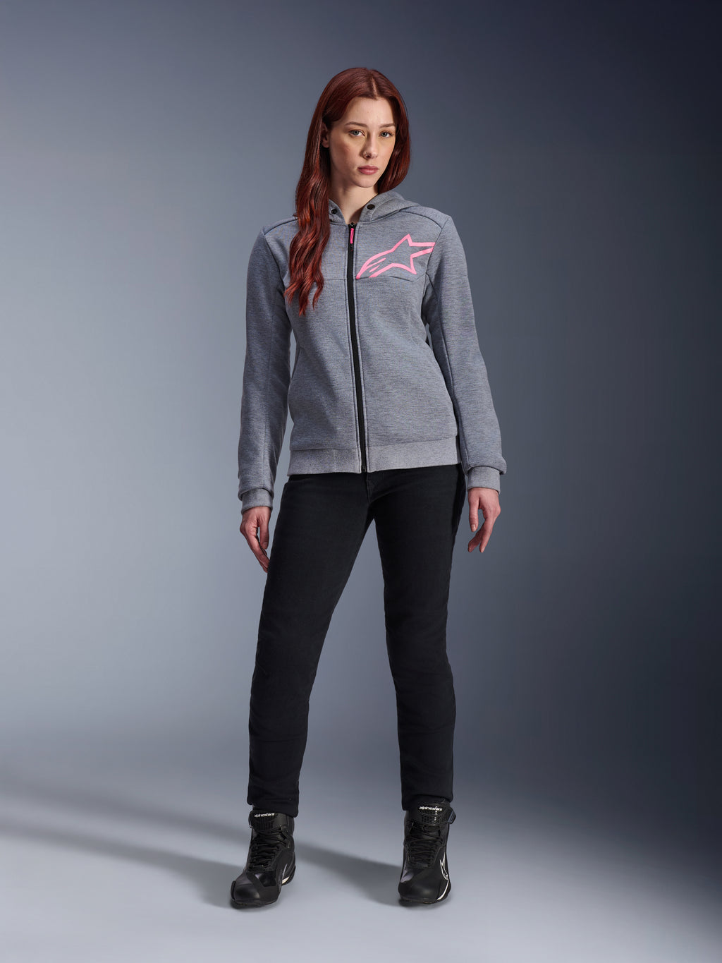 Alpinestars Stella Chrome V2 Sport Hoodie, felpa da moto con cappuccio, grigio melange e rosa fluo, indossata da una donna con pantaloni neri e stivali, logo a stella Alpinestars rosa sul petto, pile tecnico leggero con vestibilità atletica casual