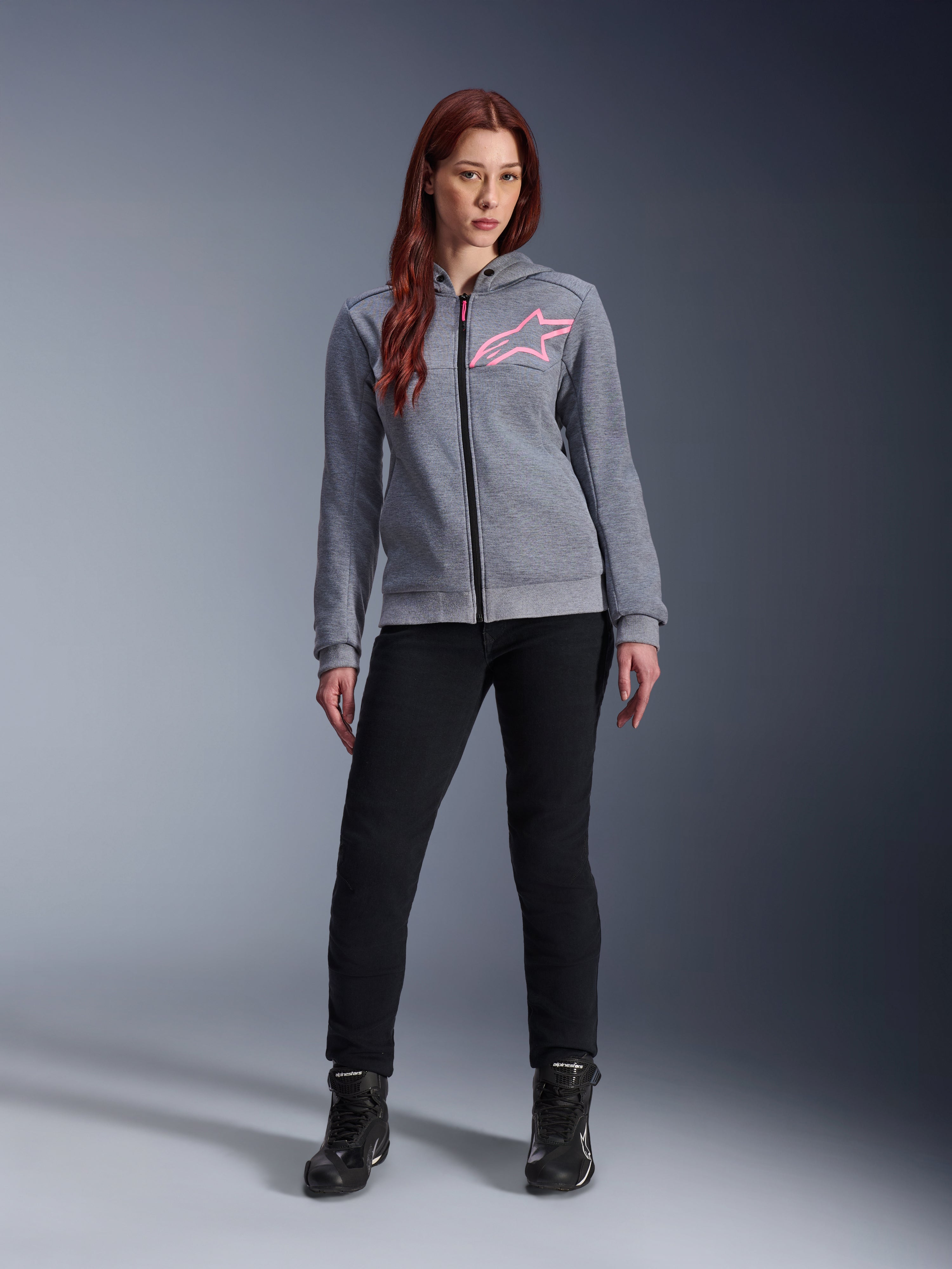 Alpinestars Stella Chrome V2 Sport Hoodie, felpa da moto con cappuccio, grigio melange e rosa fluo, indossata da una donna con pantaloni neri e stivali, logo a stella Alpinestars rosa sul petto, pile tecnico leggero con vestibilità atletica casual