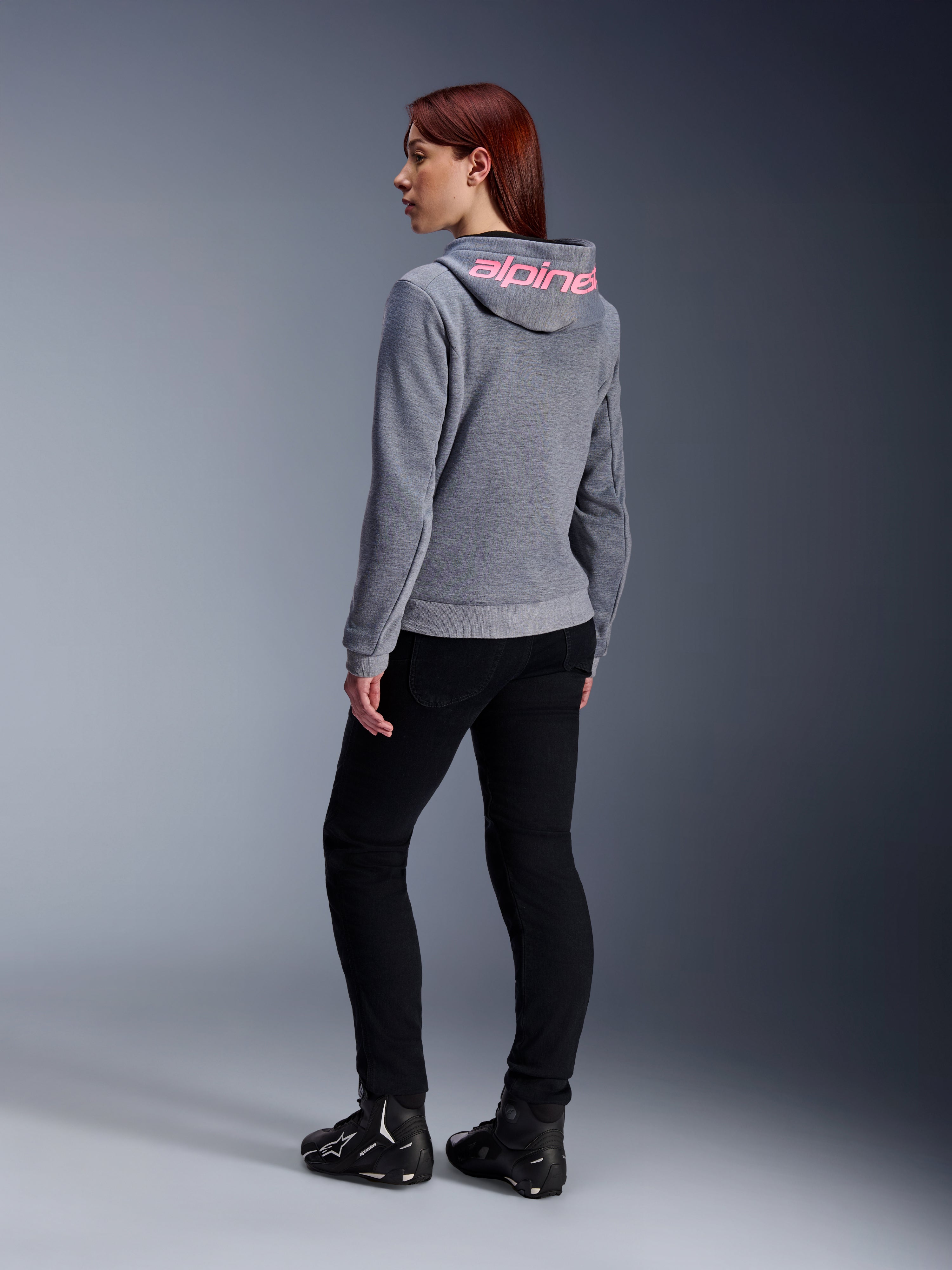 Alpinestars Stella Chrome V2 Sport Hoodie, felpa da moto da donna, grigio mélange con logo rosa fluo sul cappuccio, vista posteriore, abbinata a pantaloni neri e scarpe tecniche da guida, stile urban casual con protezione da moto