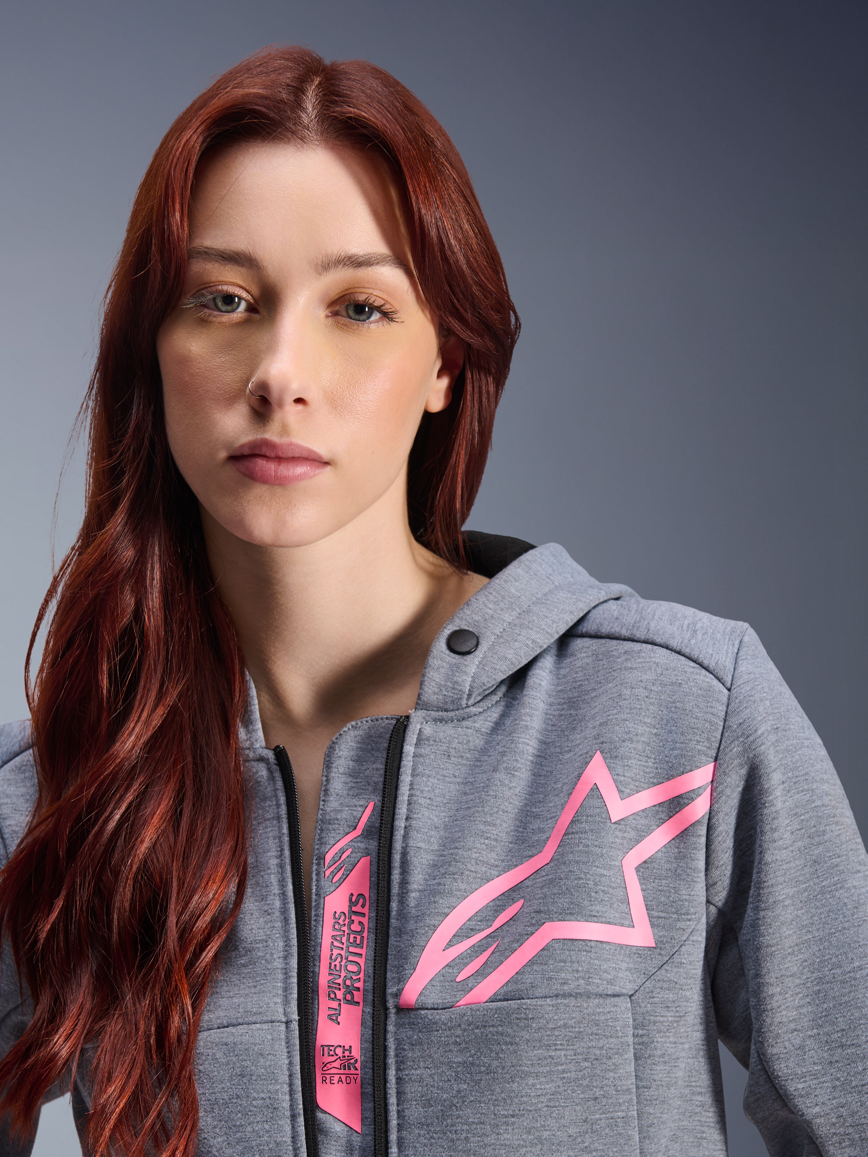Donna che indossa la felpa Alpinestars Stella Chrome V2 Sport Hoodie, Gray Heather Pink Fluo, primo piano che mostra il logo a stella rosa e il marchio Tech-Air Ready, abbigliamento moto urbano