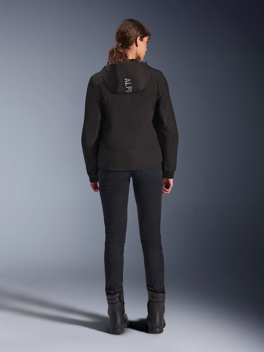 Alpinestars Woman Stella Radium Tech Hoodie, nera, vista posteriore della modella che indossa la felpa tecnica leggera a 3 strati antivento con logo Alpinestars sul cappuccio, abbinata a pantaloni e stivali neri, abbigliamento da moto urbano