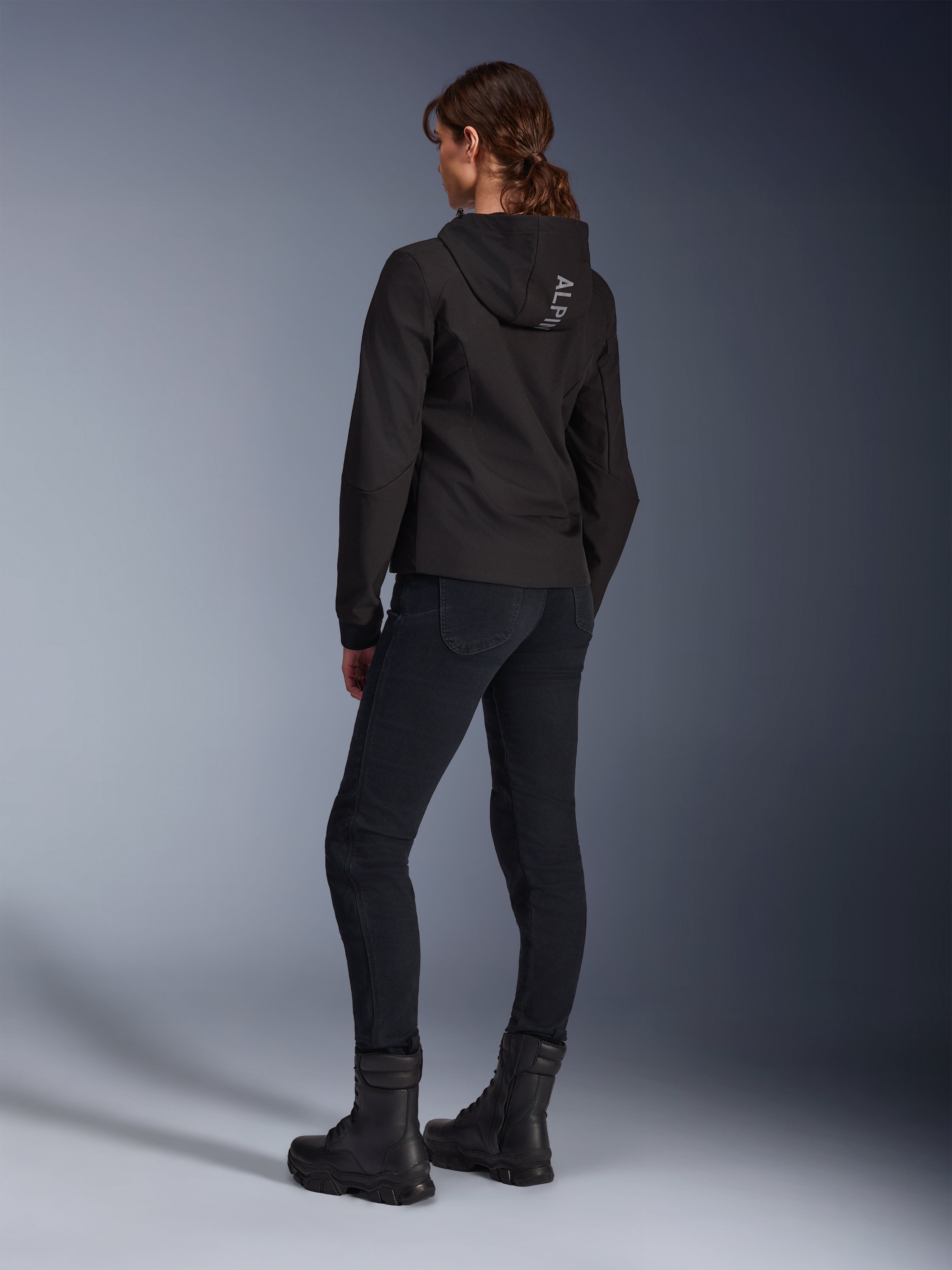 Alpinestars Stella Radium Tech Hoodie, felpa da moto urbana da donna nera, vista posteriore con logo 'ALPIN' sul cappuccio, abbinata a pantaloni tecnici da guida neri e stivali neri, guscio tecnico leggero antivento.