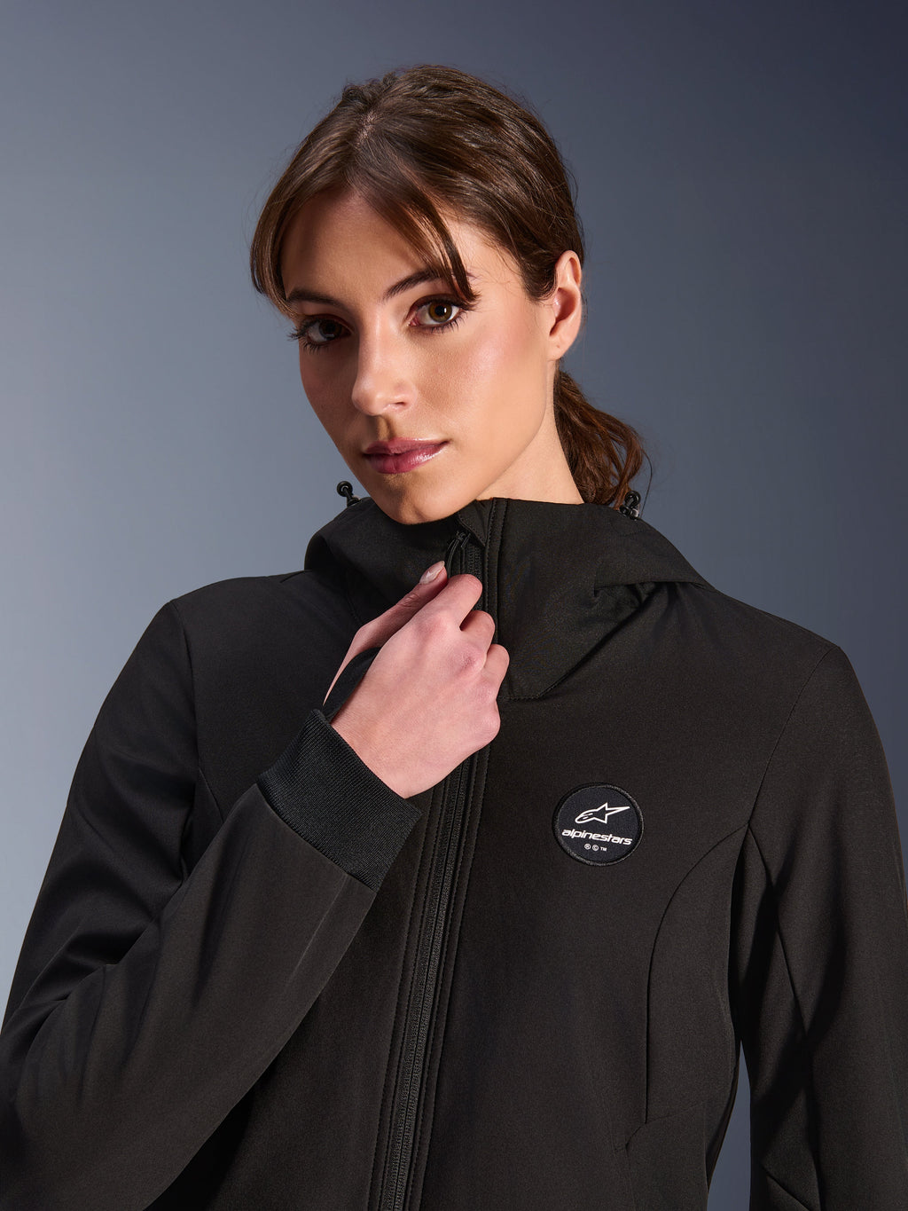 Alpinestars Stella Radium Tech Hoodie, nera, indossata da una donna che regola la cerniera frontale, con patch circolare del logo Alpinestars sul petto, polsini a coste elasticizzati e costruzione in tessuto antivento per la guida urbana.