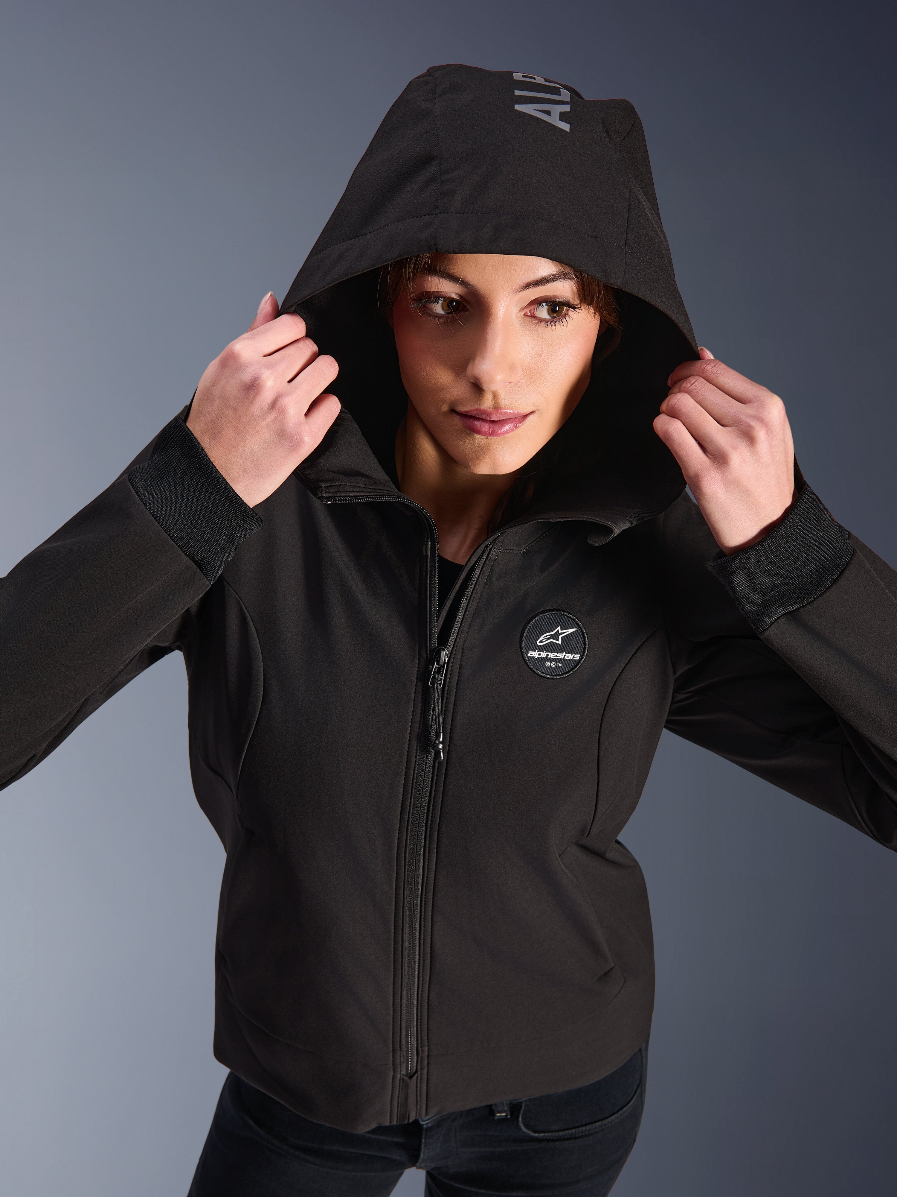 Donna che indossa la Alpinestars Stella Radium Tech Hoodie in nero, felpa da moto urbana con cappuccio alzato, caratterizzata dal logo circolare Alpinestars sul petto, polsini a coste e costruzione antivento a 3 strati