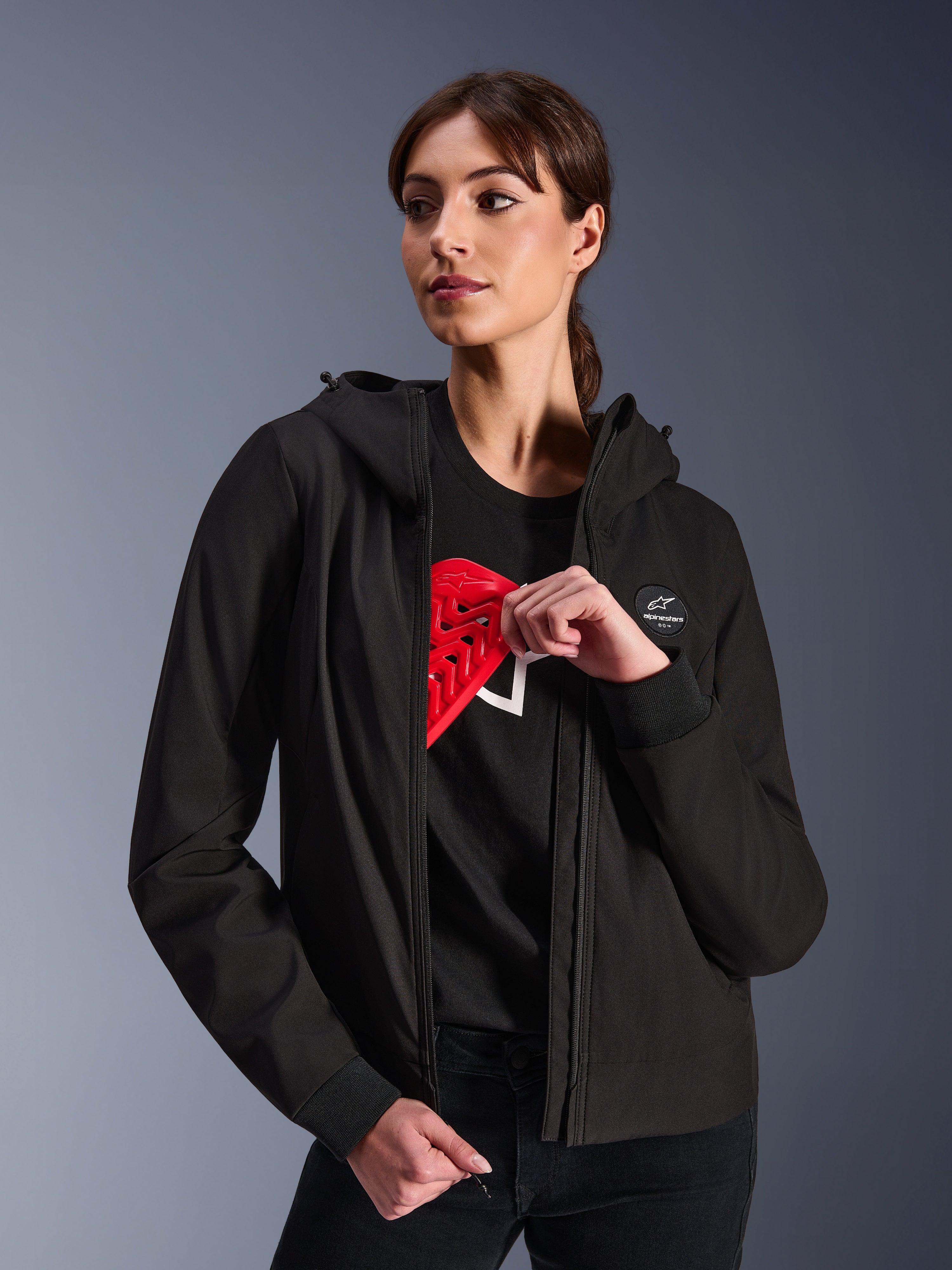 Alpinestars Stella Radium Tech Hoodie da donna, felpa da moto urbana nera, modella che mostra la protezione rossa Nucleon Flex Plus all'interno, guscio in tessuto tecnico a 3 strati leggero e antivento con polsini a coste e patch con logo sul petto, abbigliamento protettivo da moto