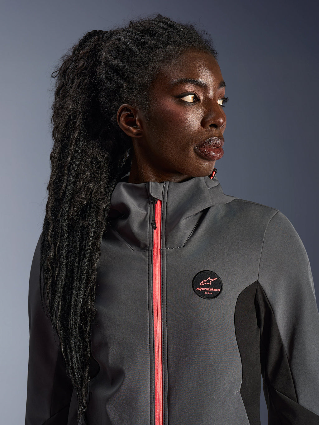 Modella che indossa la Alpinestars Stella Radium Tech Hoodie in grigio scuro e nero, con cerniera color corallo acceso, logo circolare sul petto e guscio antivento a collo alto, abbigliamento da moto urbano casual per donna