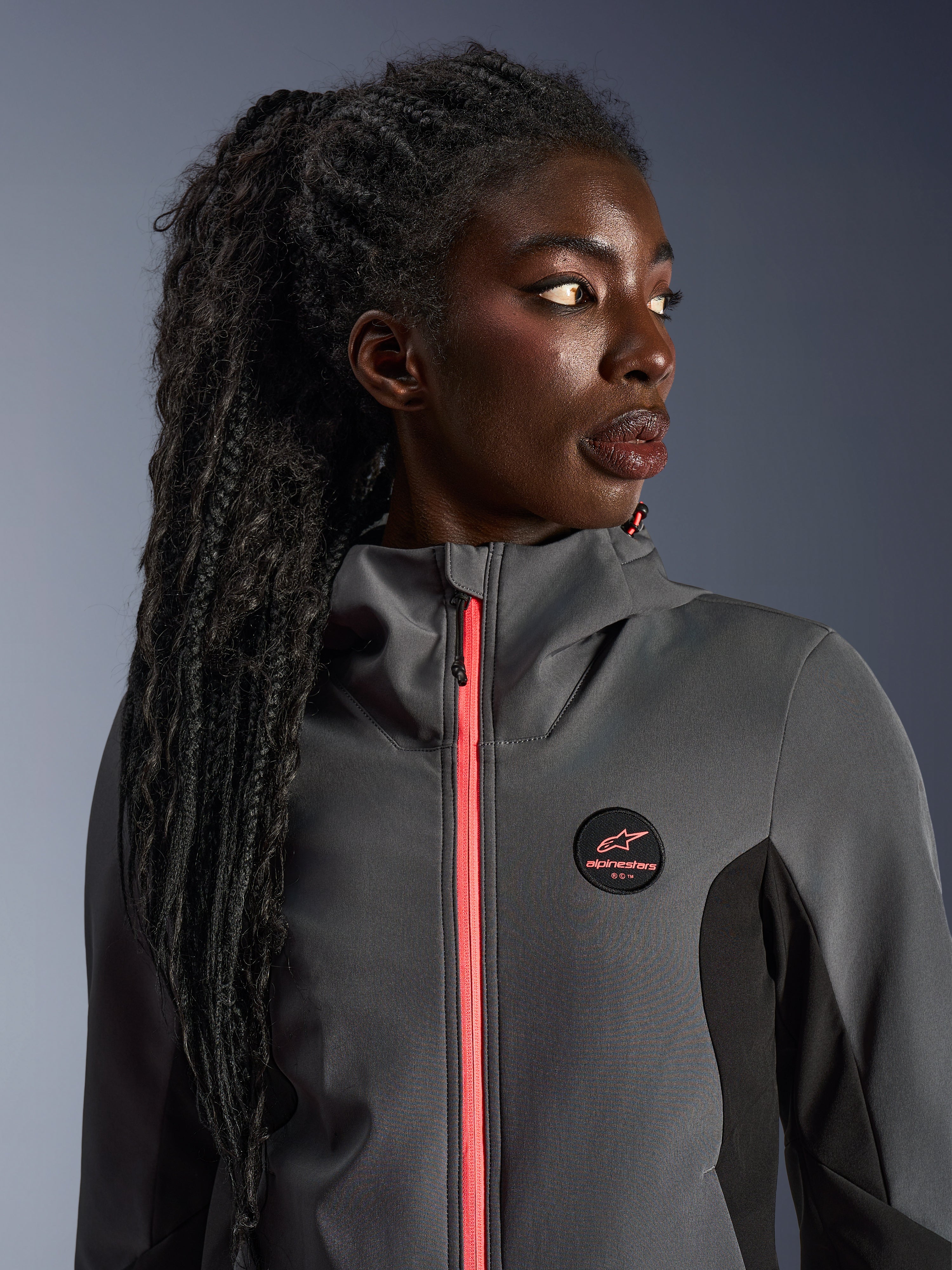 Modella che indossa la Alpinestars Stella Radium Tech Hoodie in grigio scuro e nero, con cerniera color corallo acceso, logo circolare sul petto e guscio antivento a collo alto, abbigliamento da moto urbano casual per donna