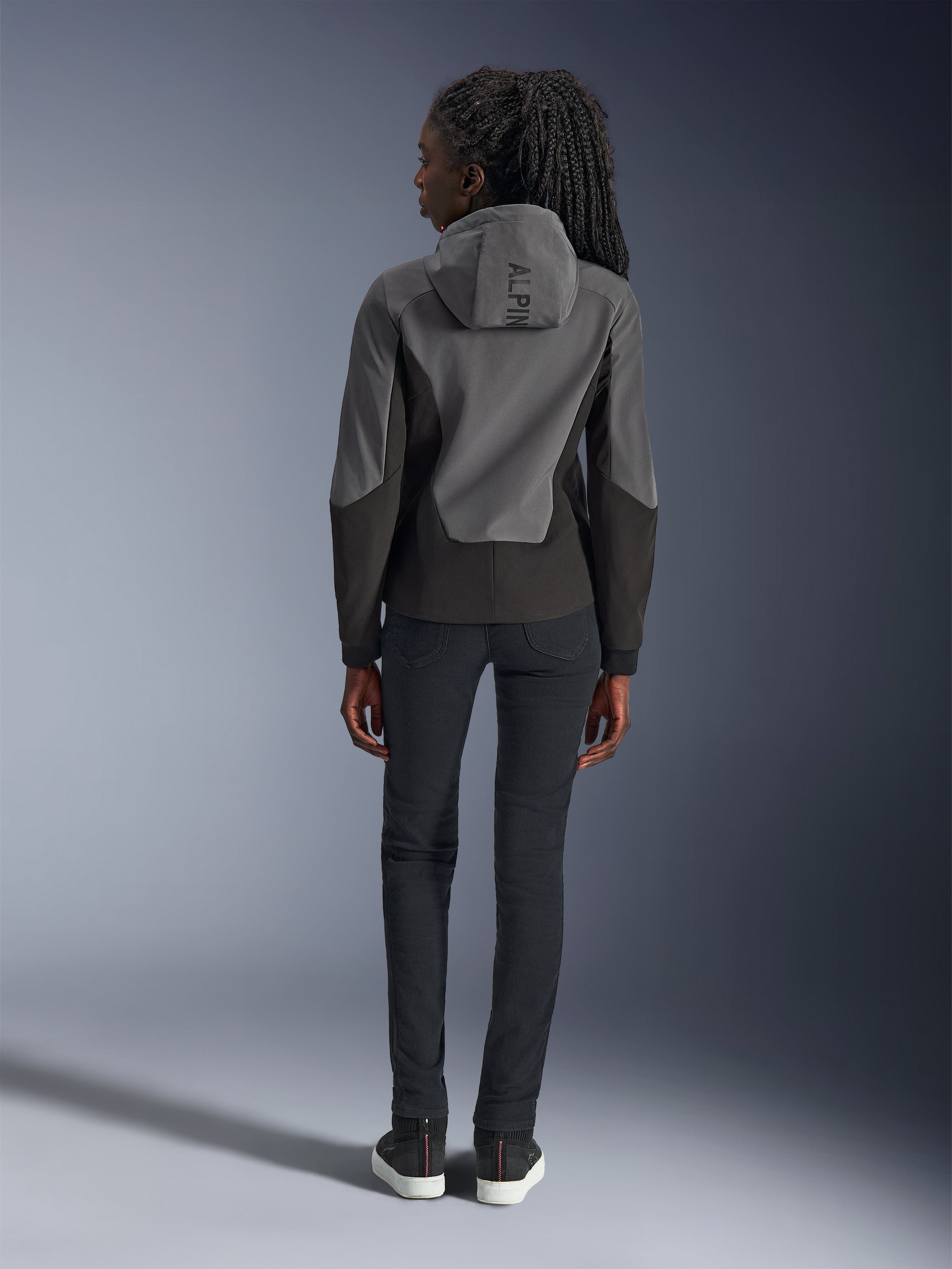 Donna che indossa la felpa Alpinestars Stella Radium Tech Hoodie, grigio scuro e nero, vista posteriore con marchio 'ALPIN' sul cappuccio, abbinata a pantaloni neri slim-fit e sneakers nere, abbigliamento da moto urbano