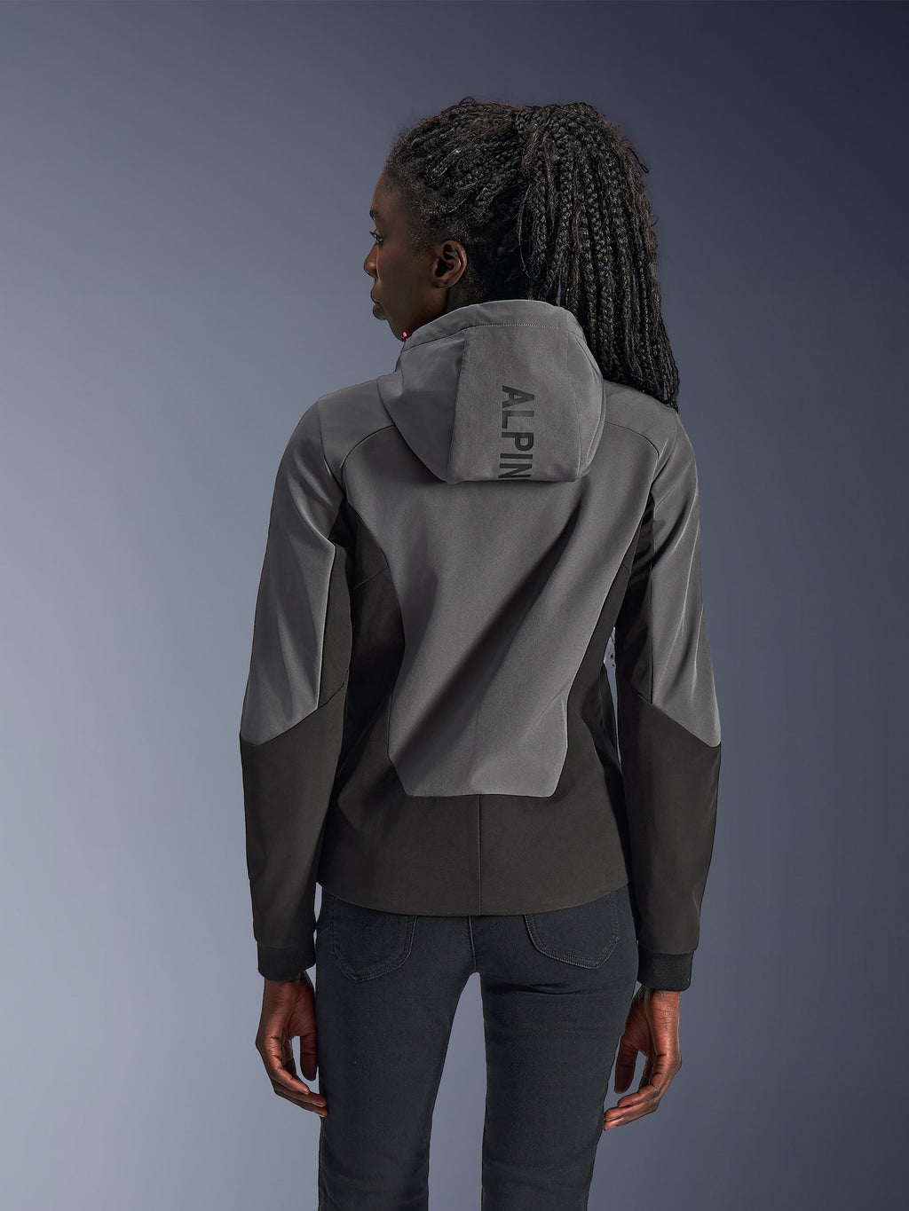Donna che indossa la felpa con cappuccio Alpinestars Stella Radium Tech, vista posteriore, Deep Gray Black, guscio antivento a 3 strati bicolore grigio e nero, marchio Alpinestars sul cappuccio, abbigliamento da moto urbano dal taglio atletico