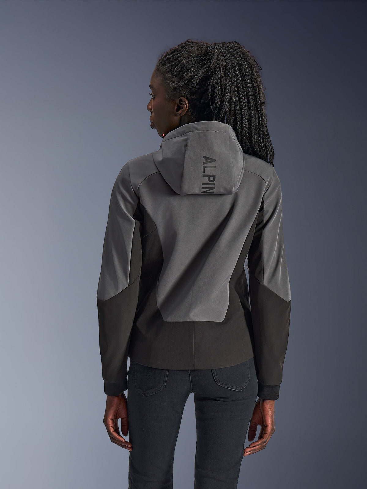 Donna che indossa la felpa con cappuccio Alpinestars Stella Radium Tech, vista posteriore, Deep Gray Black, guscio antivento a 3 strati bicolore grigio e nero, marchio Alpinestars sul cappuccio, abbigliamento da moto urbano dal taglio atletico