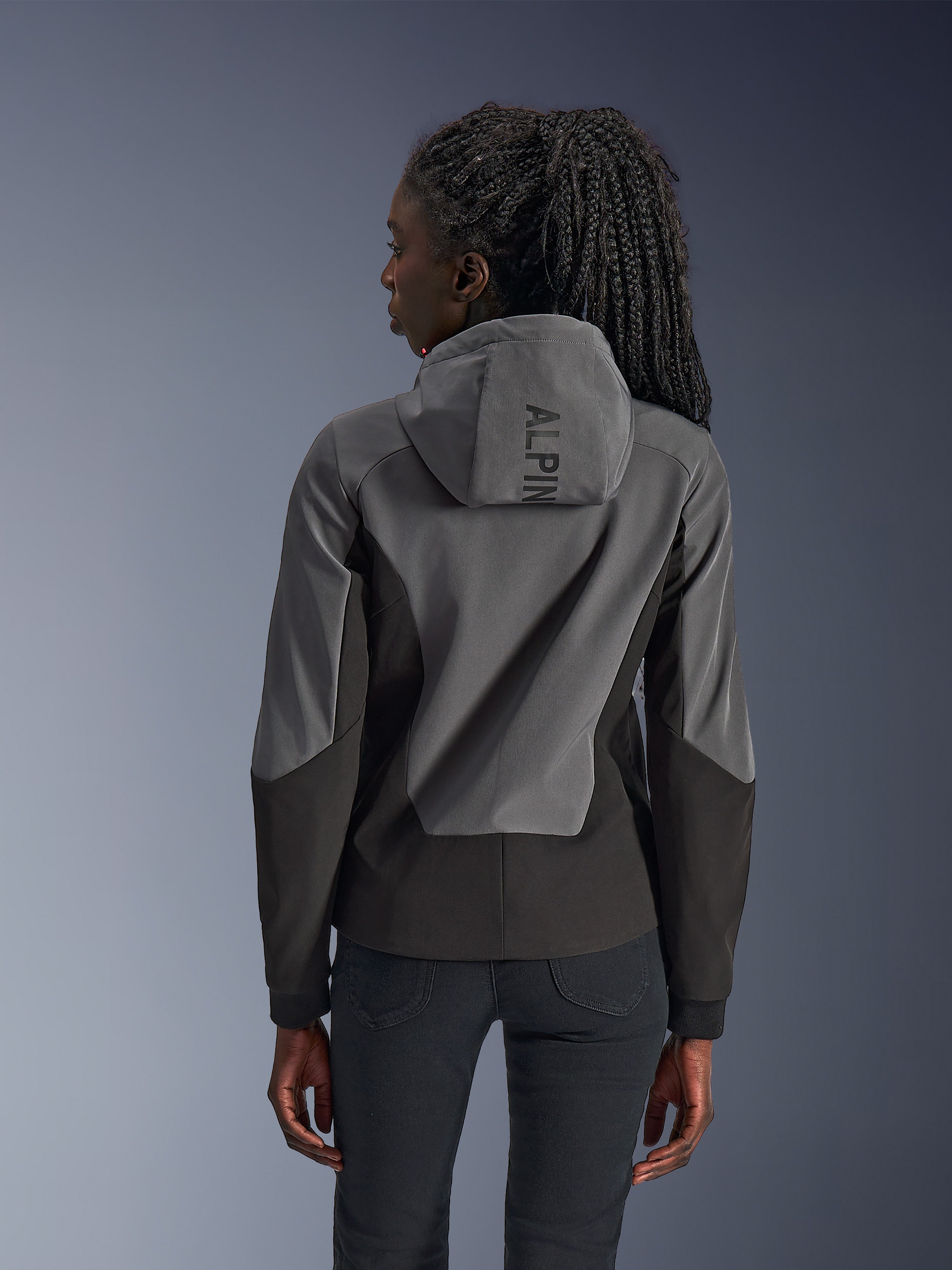 Donna che indossa la felpa con cappuccio Alpinestars Stella Radium Tech, vista posteriore, Deep Gray Black, guscio antivento a 3 strati bicolore grigio e nero, marchio Alpinestars sul cappuccio, abbigliamento da moto urbano dal taglio atletico