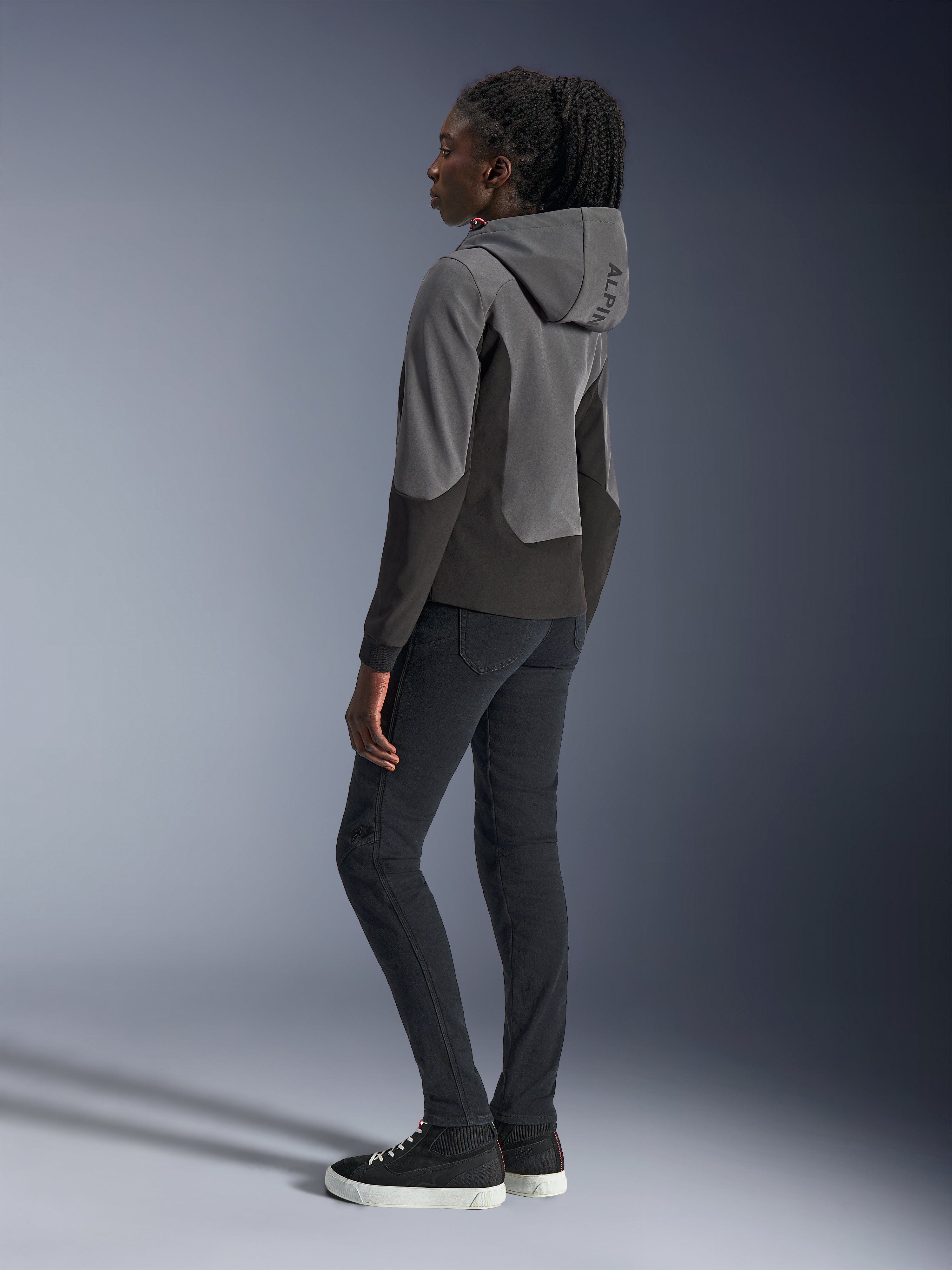 Donna che indossa la felpa con cappuccio Alpinestars Stella Radium Tech in Deep Gray e Black, abbigliamento moto urbano, vista posteriore che mostra il cappuccio con marchio Alpinestars, abbinata a pantaloni slim-fit neri e sneaker alte