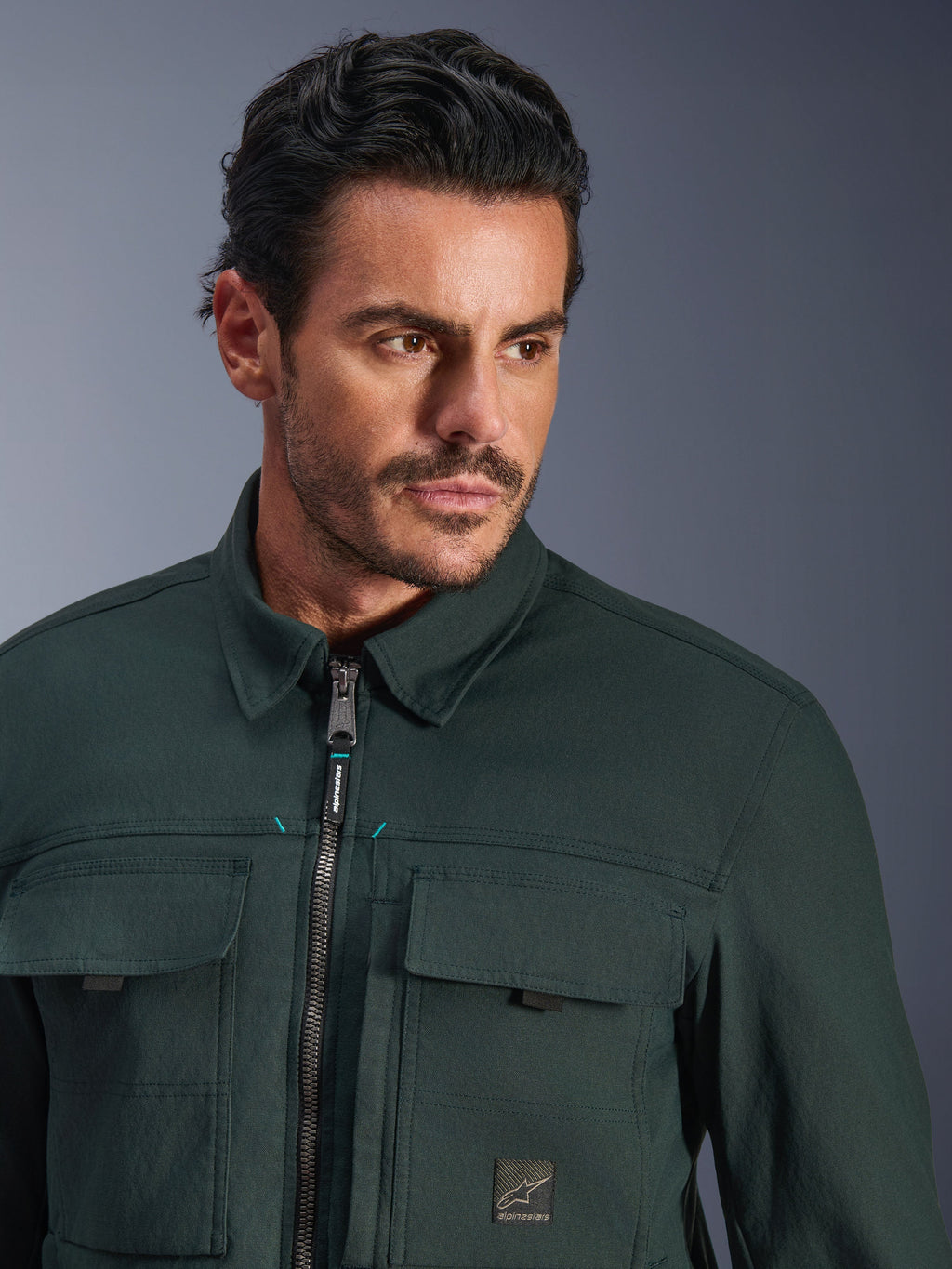 Alpinestars Bryon Water Resistant Overshirt in Dark Emerald, primo piano di un uomo che indossa la giacca da moto urban, con texture in tela cerata, zip frontale con discreti accenti teal, tasche cargo sul petto e patch con logo del marchio.