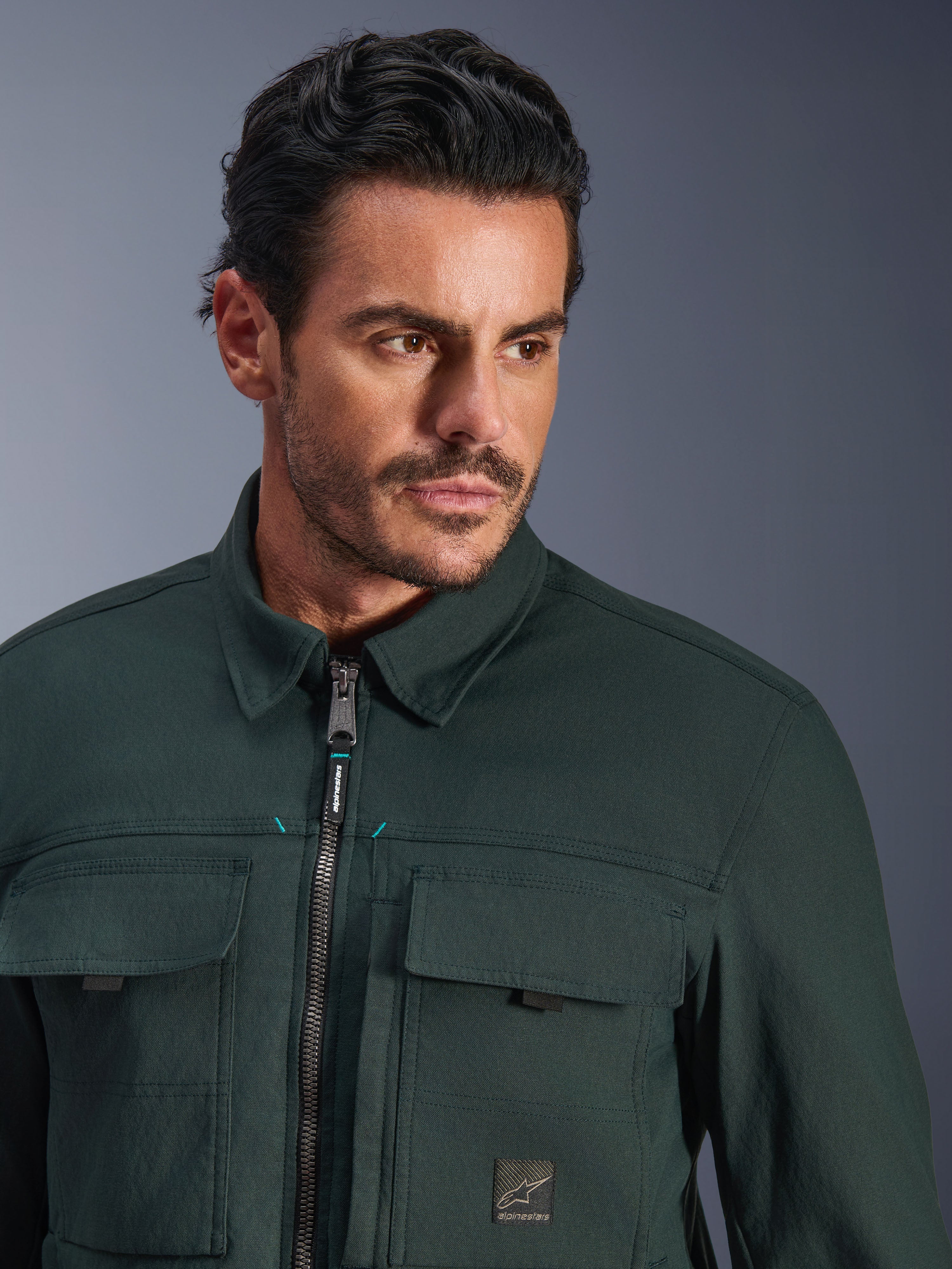 Alpinestars Bryon Water Resistant Overshirt in Dark Emerald, primo piano di un uomo che indossa la giacca da moto urban, con texture in tela cerata, zip frontale con discreti accenti teal, tasche cargo sul petto e patch con logo del marchio.