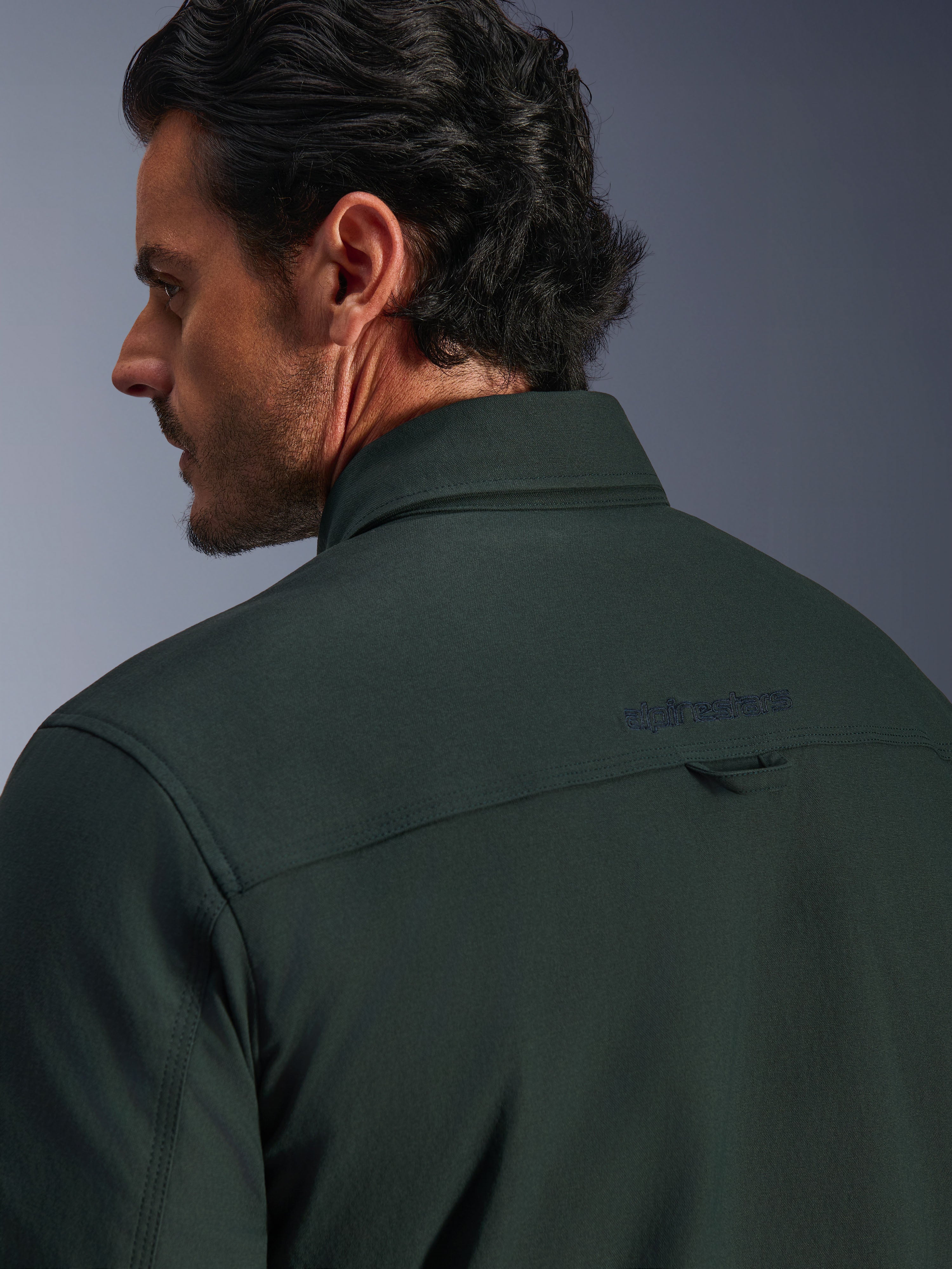 Alpinestars Bryon Water Resistant Overshirt in verde Dark Emerald, primo piano della parte superiore della schiena e del colletto su un modello, con logo Alpinestars ricamato e un piccolo passante in tessuto, in tela cerata testurizzata resistente all'acqua per uso urbano.