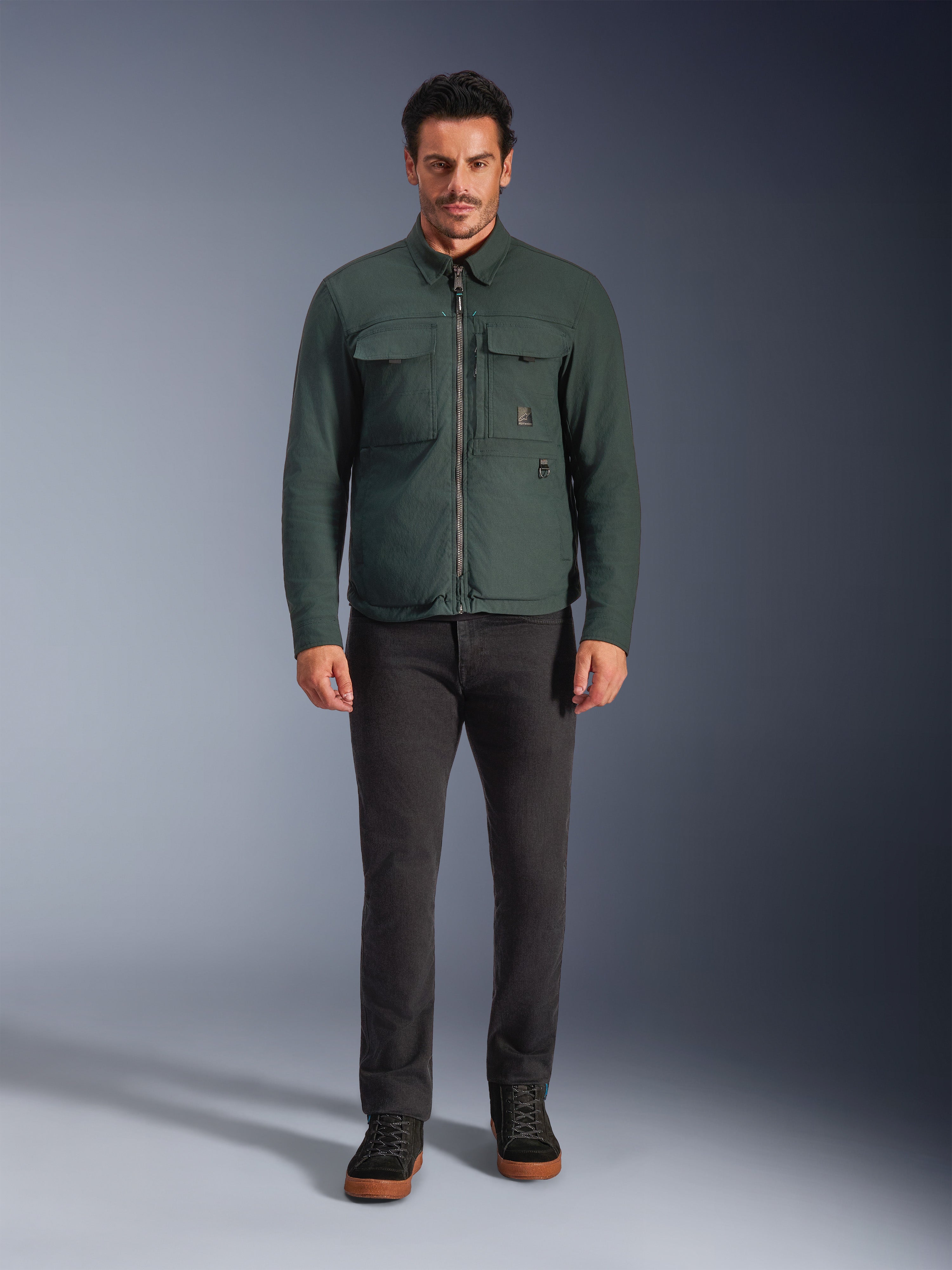 Alpinestars Bryon WR Overshirt, giacca da moto resistente all'acqua, smeraldo scuro, indossata con pantaloni neri e stivali neri, caratterizzata da una costruzione in tela cerata, tasche cargo sul petto e un design urbano moderno.
