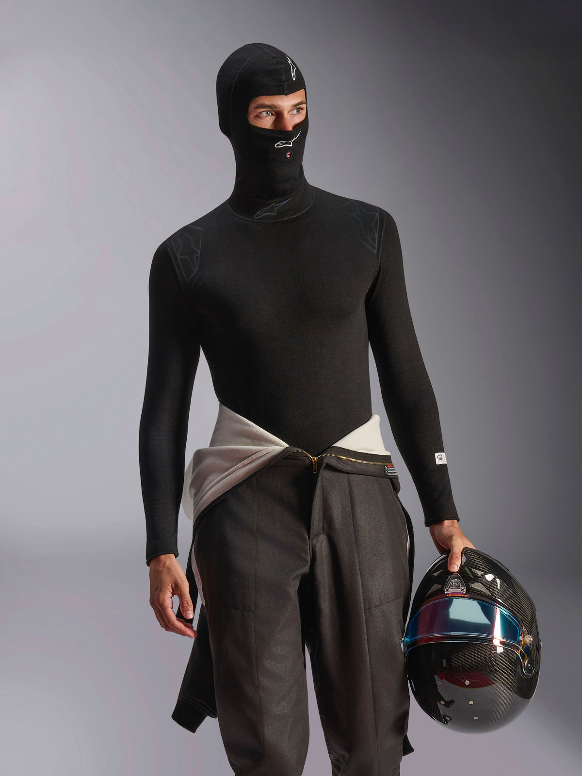 Modello con passamontagna Alpinestars ZX Evo V3 nero, con loghi bianchi su fronte e mento, abbinato a un base layer nero a maniche lunghe e un casco da corsa in fibra di carbonio, abbigliamento ignifugo professionale per corse automobilistiche.
