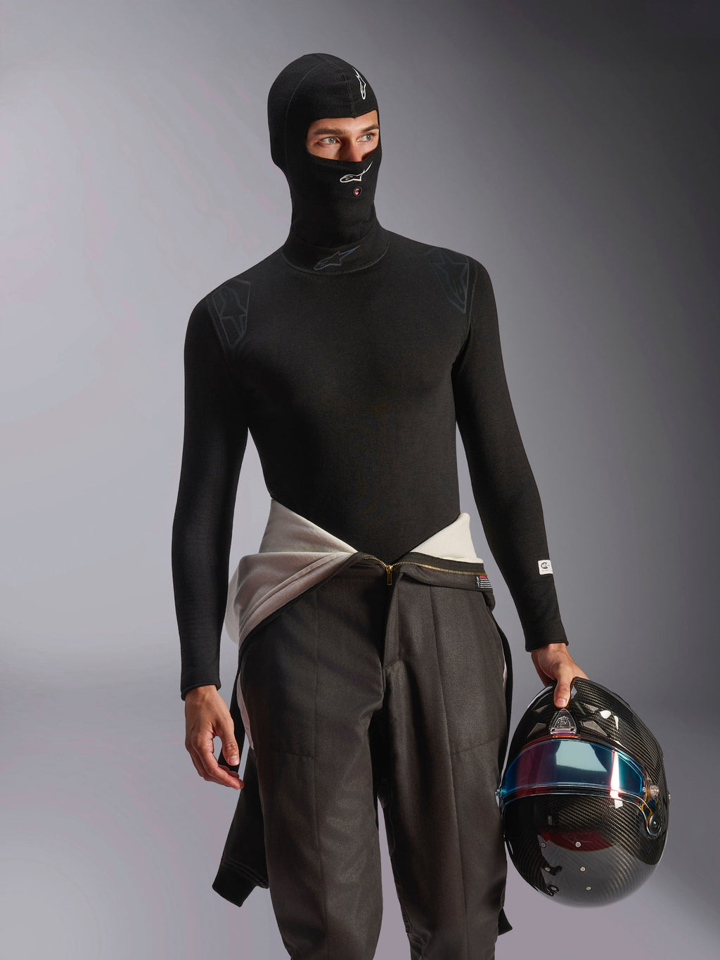 Modello con passamontagna Alpinestars ZX Evo V3 nero, con loghi bianchi su fronte e mento, abbinato a un base layer nero a maniche lunghe e un casco da corsa in fibra di carbonio, abbigliamento ignifugo professionale per corse automobilistiche.