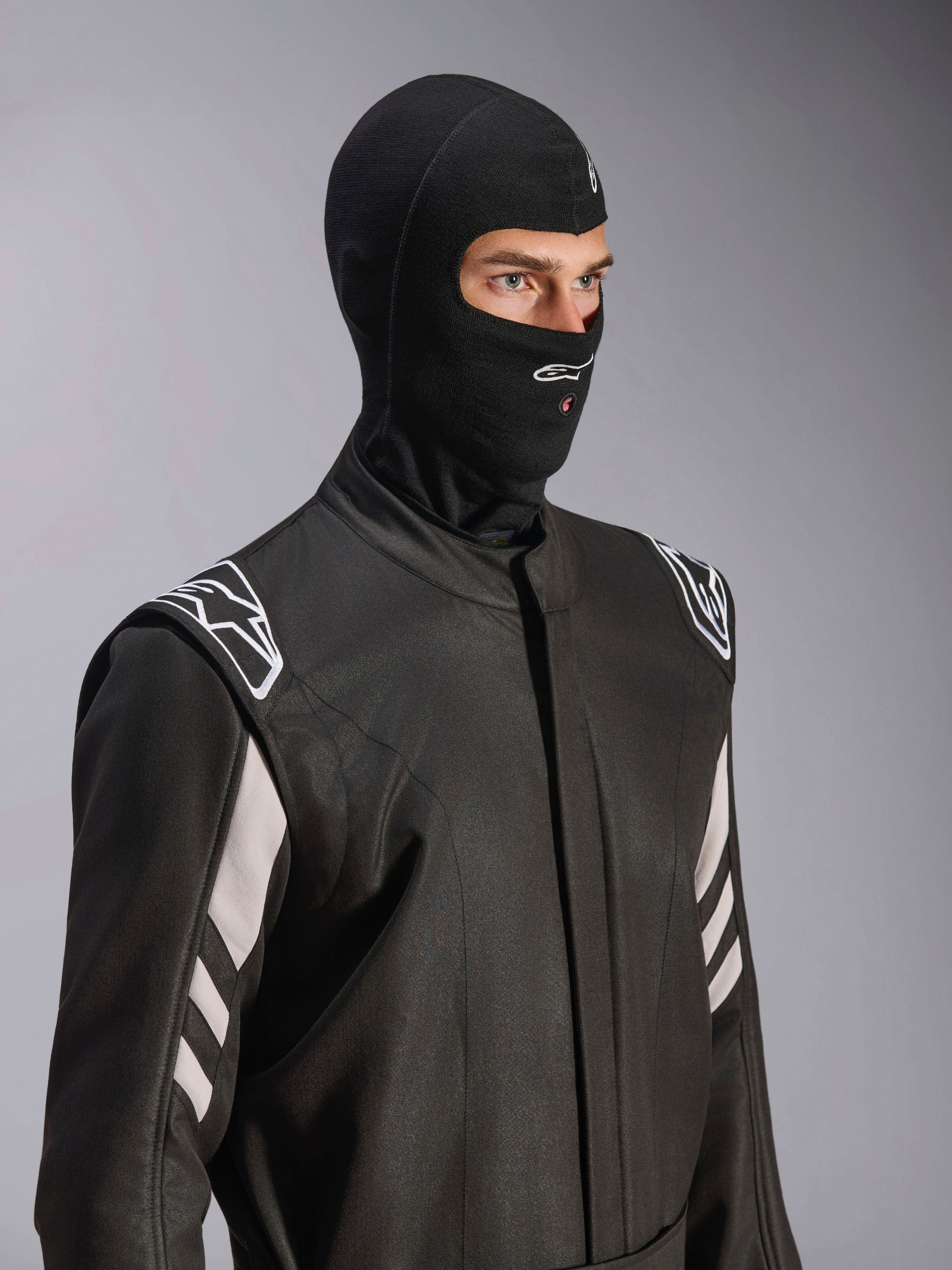 Alpinestars ZX Evo V3 Balaclava, nero, passamontagna ignifugo per corse automobilistiche, indossato da un modello in tuta da corsa nera con accenti bianchi, logo Alpinestars sul mento, apertura per il tubo di idratazione, equipaggiamento protettivo professionale per motorsport