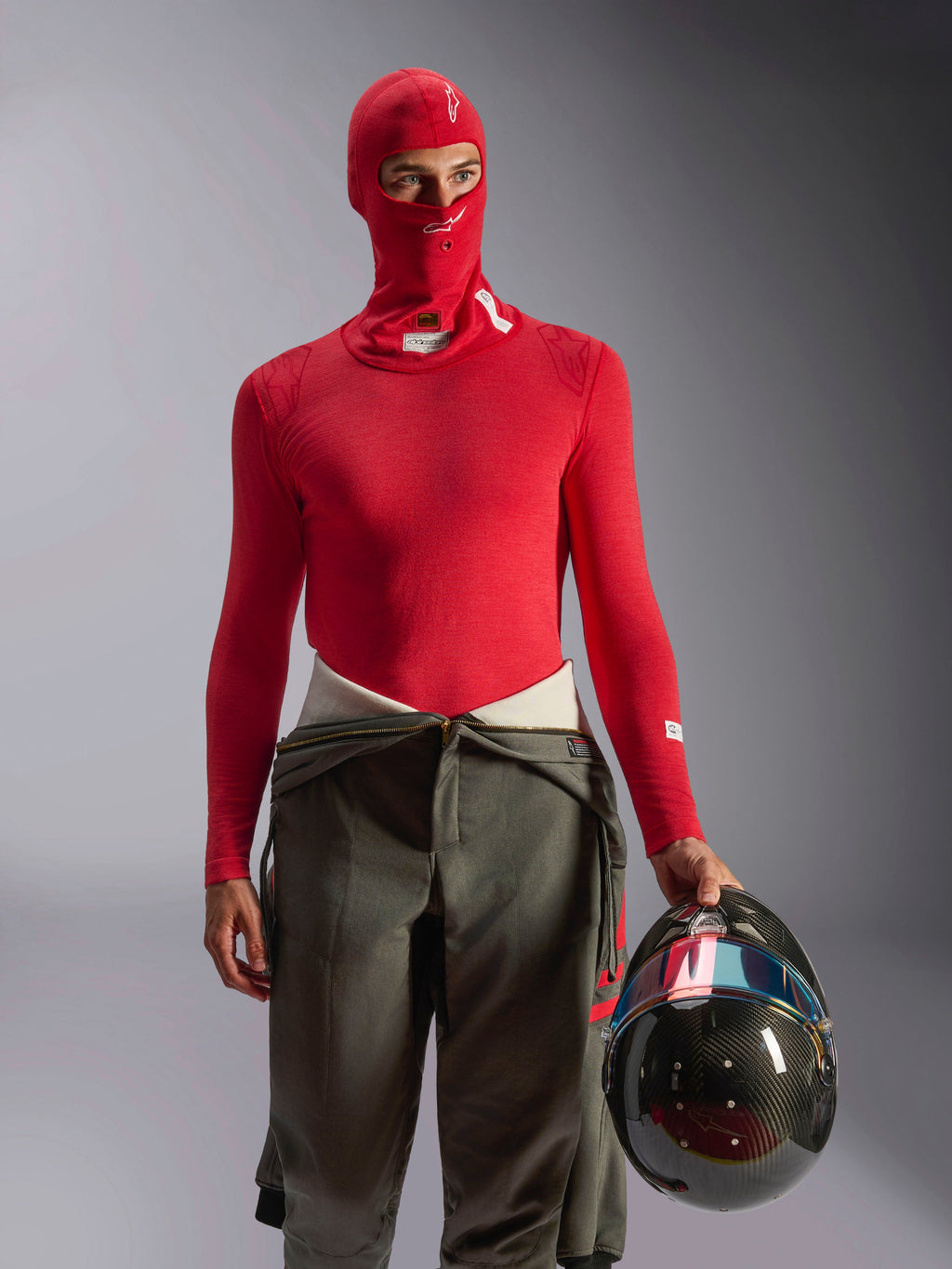 Alpinestars ZX Evo V3 Balaclava in rosso, strato di base per automobilismo indossato da un pilota con maglia a maniche lunghe rossa abbinata e pantaloni da corsa verde oliva, con in mano un casco in fibra di carbonio, abbigliamento professionale ignifugo per motorsport