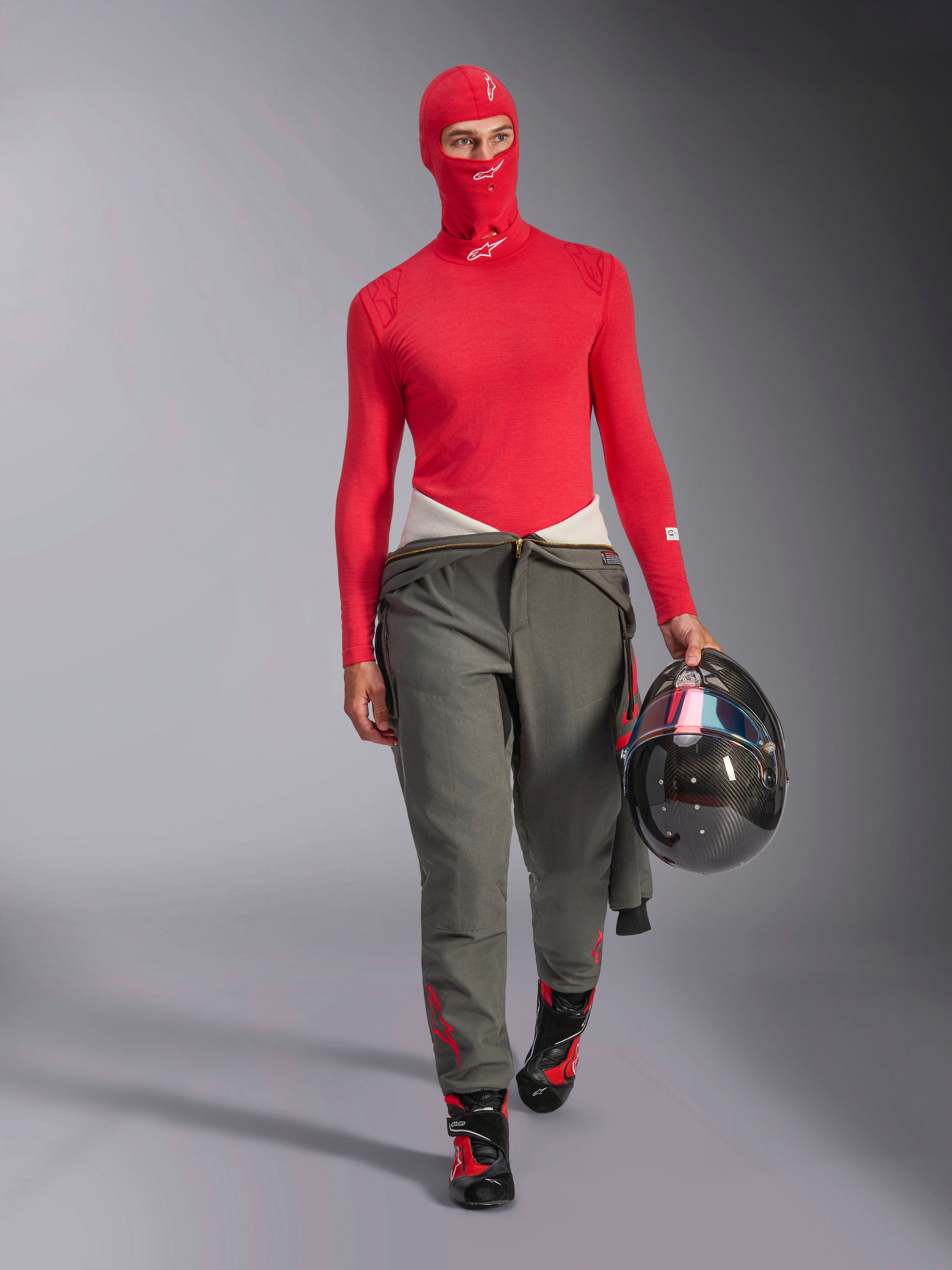 Pilota da corsa che indossa il passamontagna Alpinestars ZX Evo V3 in rosso, base layer coordinato a maniche lunghe rosso, pantaloni da gara grigi, stivali da racing neri e rossi, con casco in fibra di carbonio, set professionale di abbigliamento per corse automobilistiche.