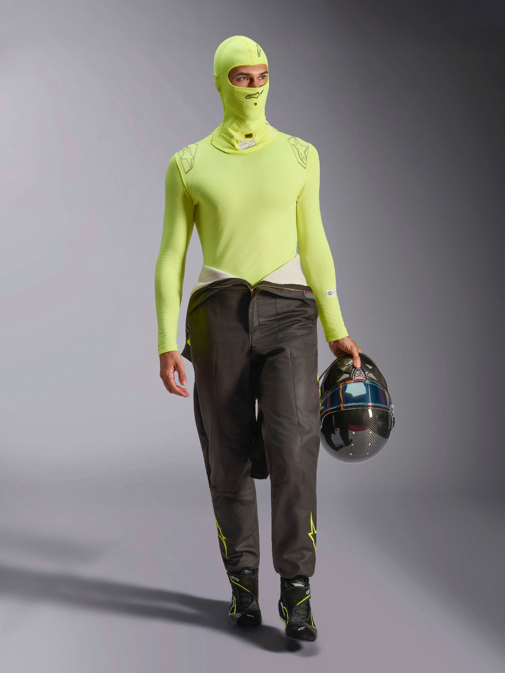 Pilota di auto da corsa con passamontagna Alpinestars ZX Evo V3 in giallo fluo, maglia base layer a maniche lunghe coordinata, pantaloni da corsa scuri e stivali da gara neri, tiene un casco in fibra di carbonio, abbigliamento professionale da competizione