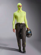 Pilota di auto da corsa con passamontagna Alpinestars ZX Evo V3 in giallo fluo, maglia base layer a maniche lunghe coordinata, pantaloni da corsa scuri e stivali da gara neri, tiene un casco in fibra di carbonio, abbigliamento professionale da competizione