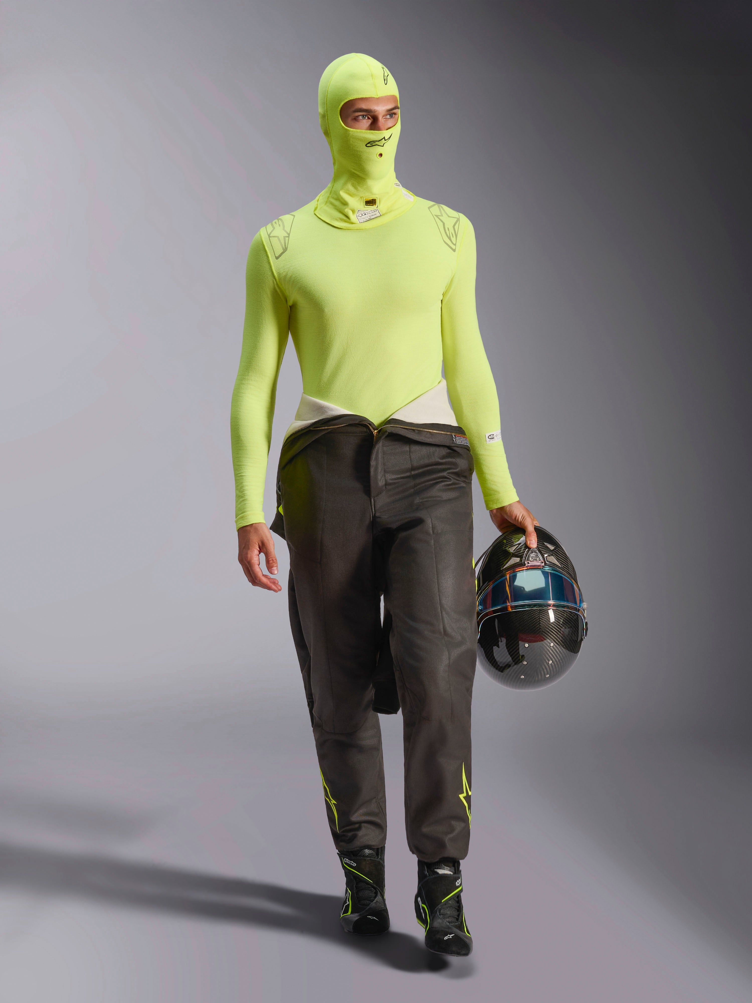 Pilota di auto da corsa con passamontagna Alpinestars ZX Evo V3 in giallo fluo, maglia base layer a maniche lunghe coordinata, pantaloni da corsa scuri e stivali da gara neri, tiene un casco in fibra di carbonio, abbigliamento professionale da competizione