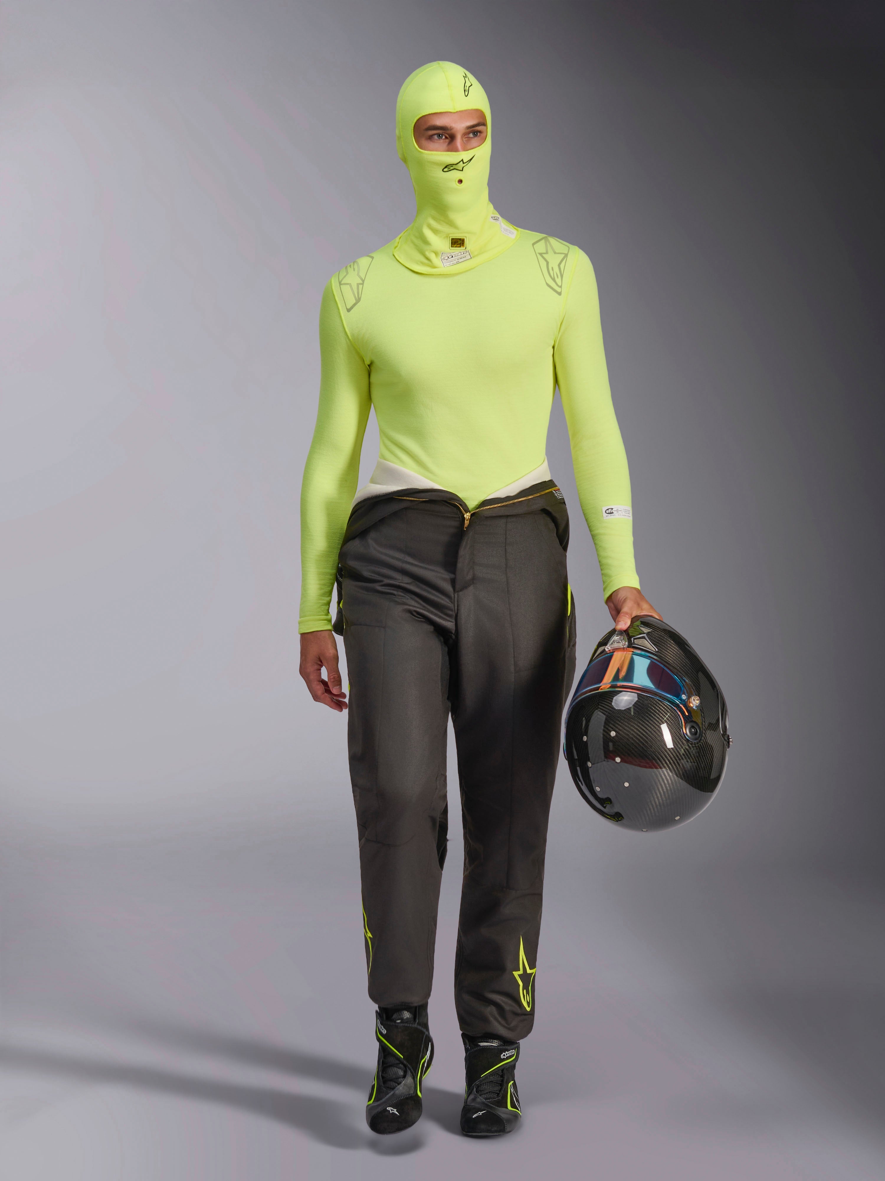 Alpinestars ZX Evo V3 Balaclava in Yellow Fluo, indossato con maglia base layer a maniche lunghe coordinata e pantaloni da corsa grigio scuro, modello maschile con in mano un casco in fibra di carbonio, attrezzatura professionale per corse automobilistiche