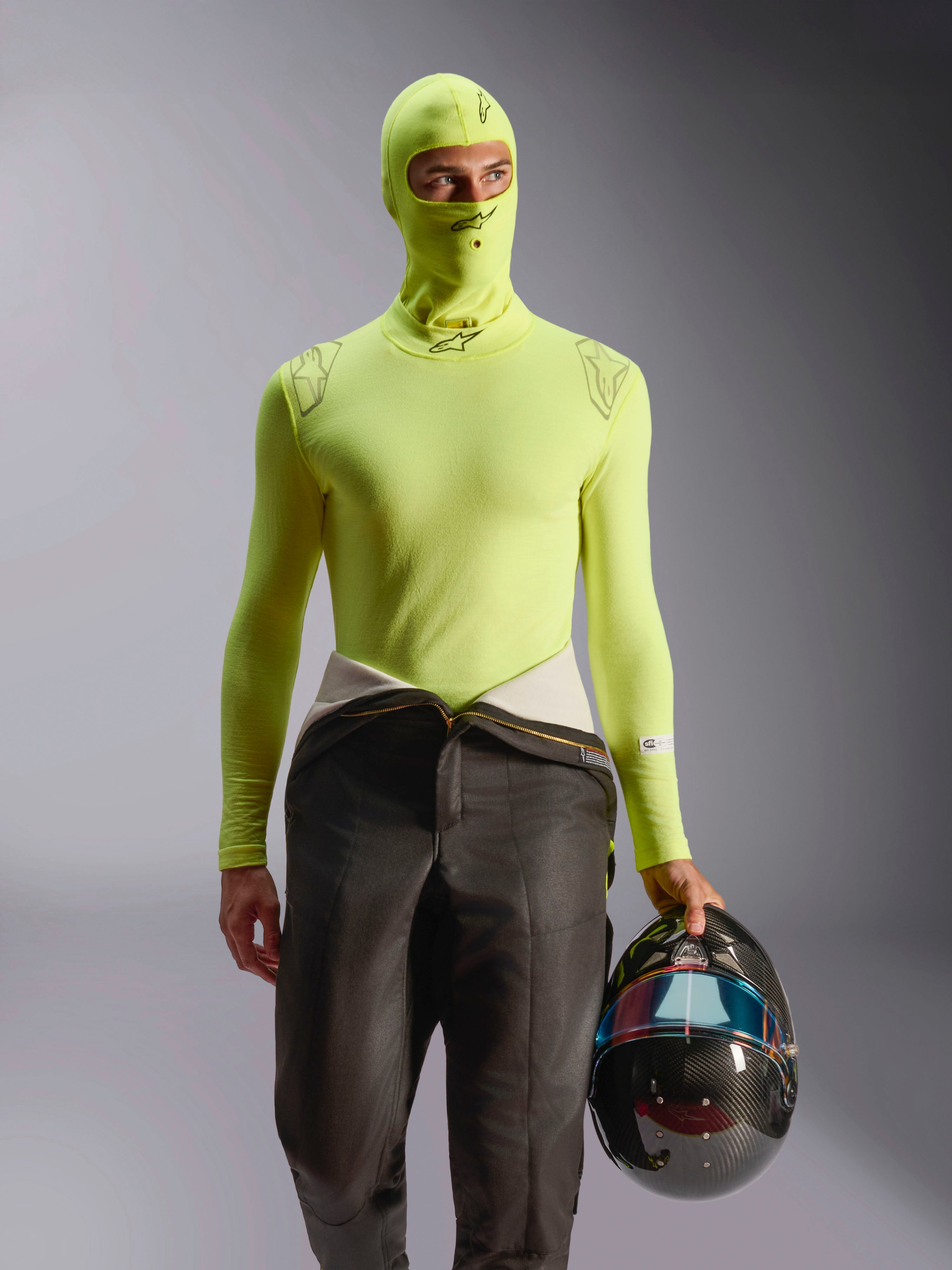 Alpinestars ZX Evo V3 Balaclava, passamontagna per corse automobilistiche, Giallo Fluo, indossato con maglia a maniche lunghe coordinata e pantaloni della tuta da corsa, modello che tiene un casco in fibra di carbonio con visiera iridescente, abbigliamento professionale ignifugo da motorsport