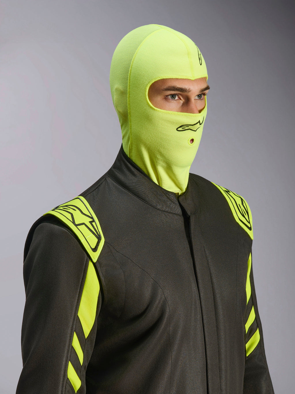 Modello che indossa il passamontagna Alpinestars ZX Evo V3 in Yellow Fluo, sottocasco da gara resistente alle fiamme, abbinato a una tuta da auto grigio scuro con accenti giallo neon, logo Alpinestars nero e apertura per tubo di idratazione, equipaggiamento protettivo professionale per motorsport.