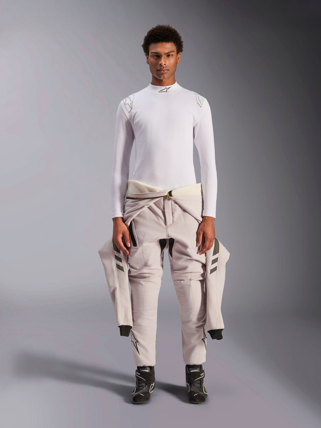 Alpinestars ZX Evo V3 Top, a maniche lunghe, bianco, base layer per corse automobilistiche indossato con una tuta da gara chiara legata in vita e stivali da corsa neri, con collo a lupetto con logo e branding sulle spalle, intimo professionale ignifugo per motorsport
